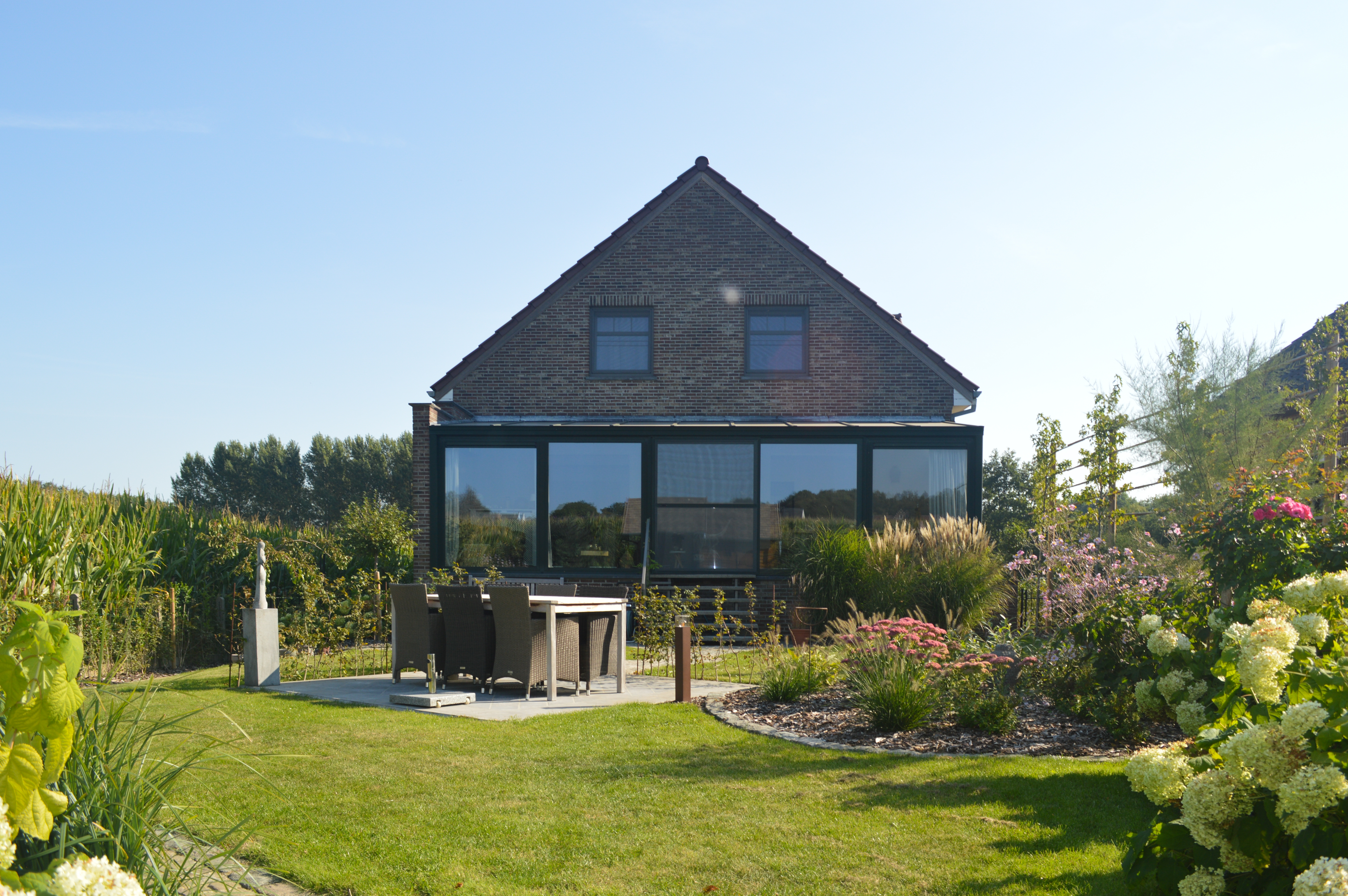 Achterzijde van een open bebouwing met gerenoveerde aanbouw, grote schuiframen met kwartsgrijze NERF-profielen en een schuifvliegenraam, tuin en terras.