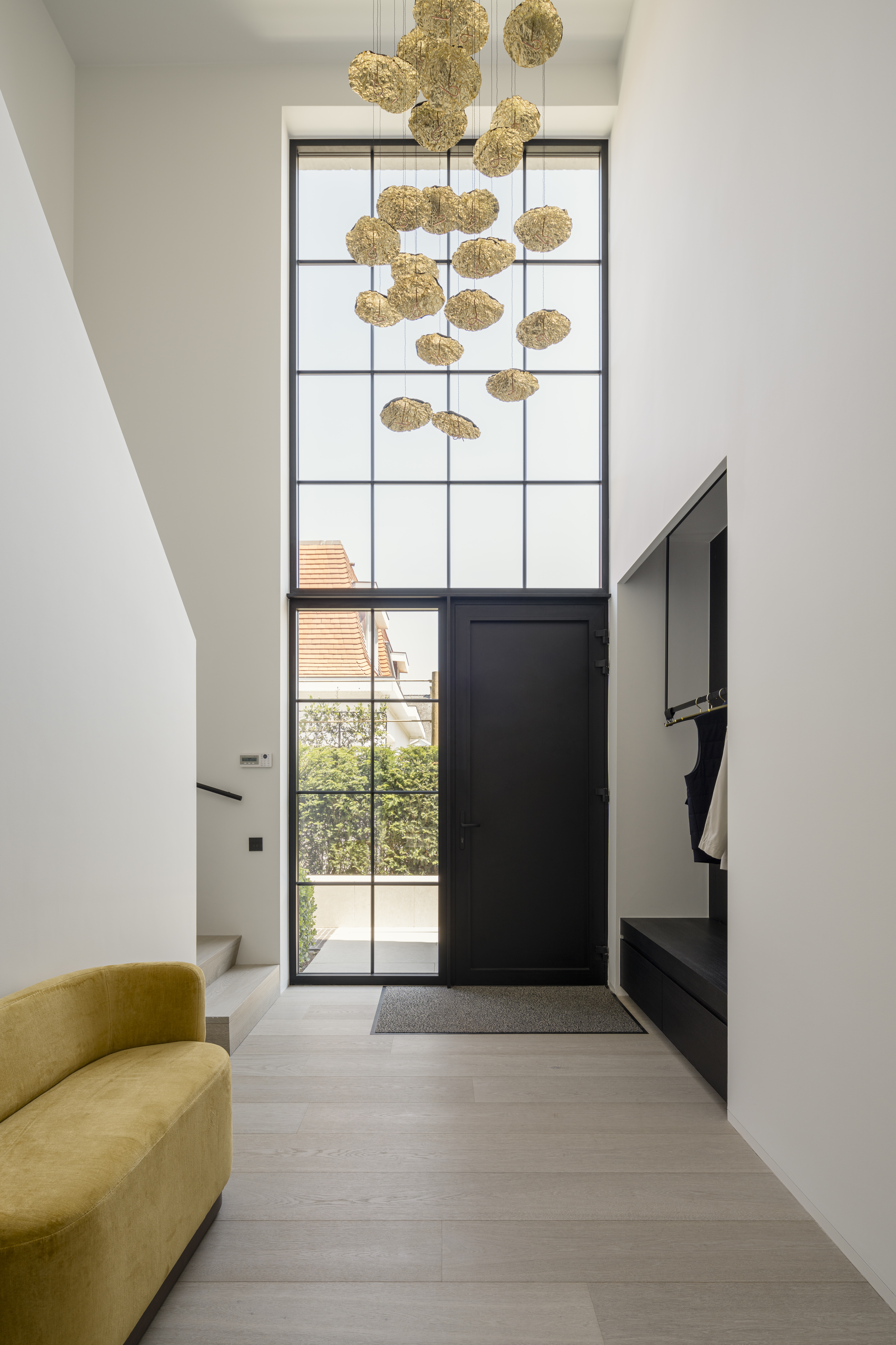 Moderne inkomhal met een hoge zwarte glazen voordeur, een lichte vloer en een hanglamp met goudkleurige bollen.