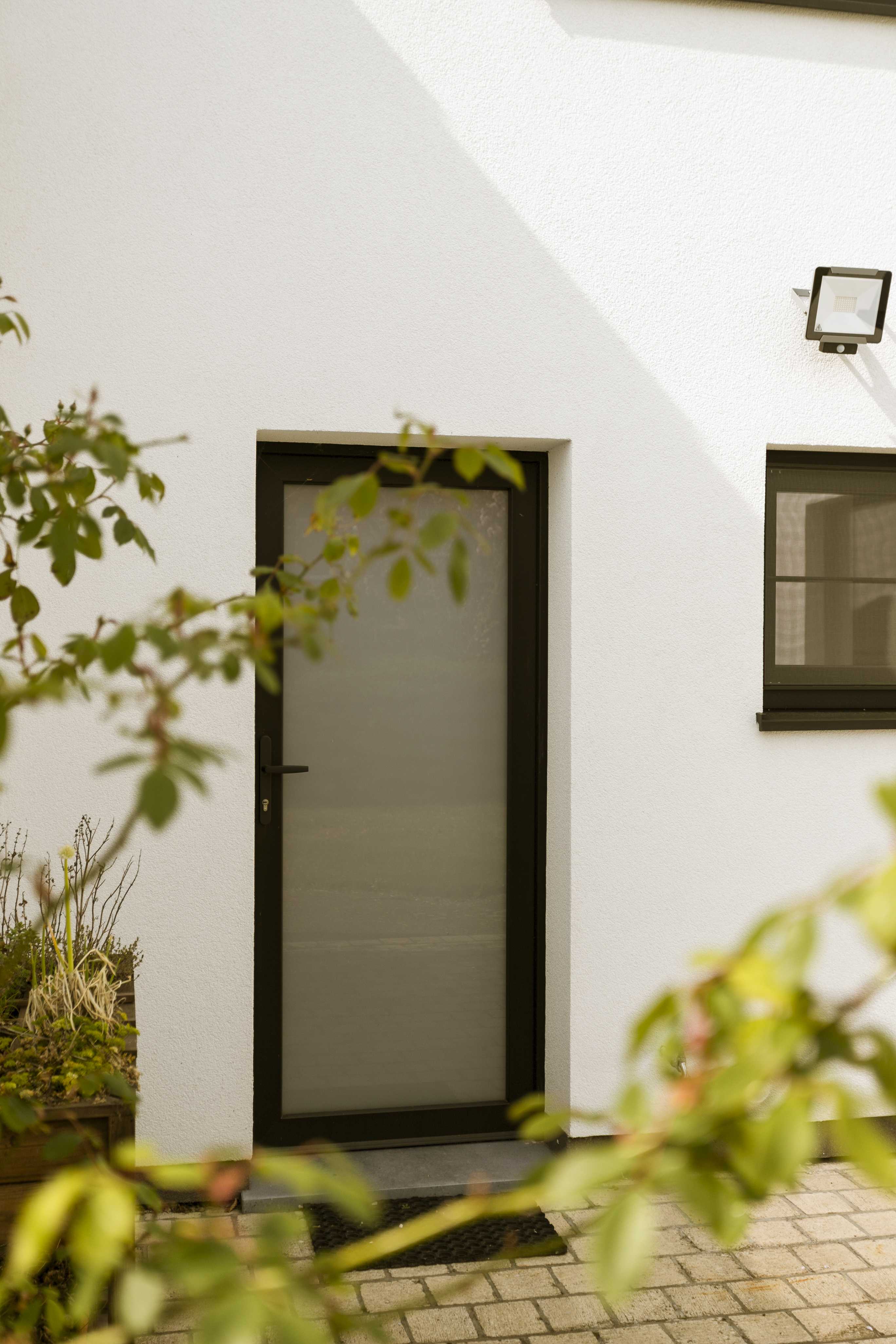 Witte gevel van een vrijstaande woning met een matzwarte, beglaasde Sablé PVC Koncept-deur, een raam en een betegelde inkom.