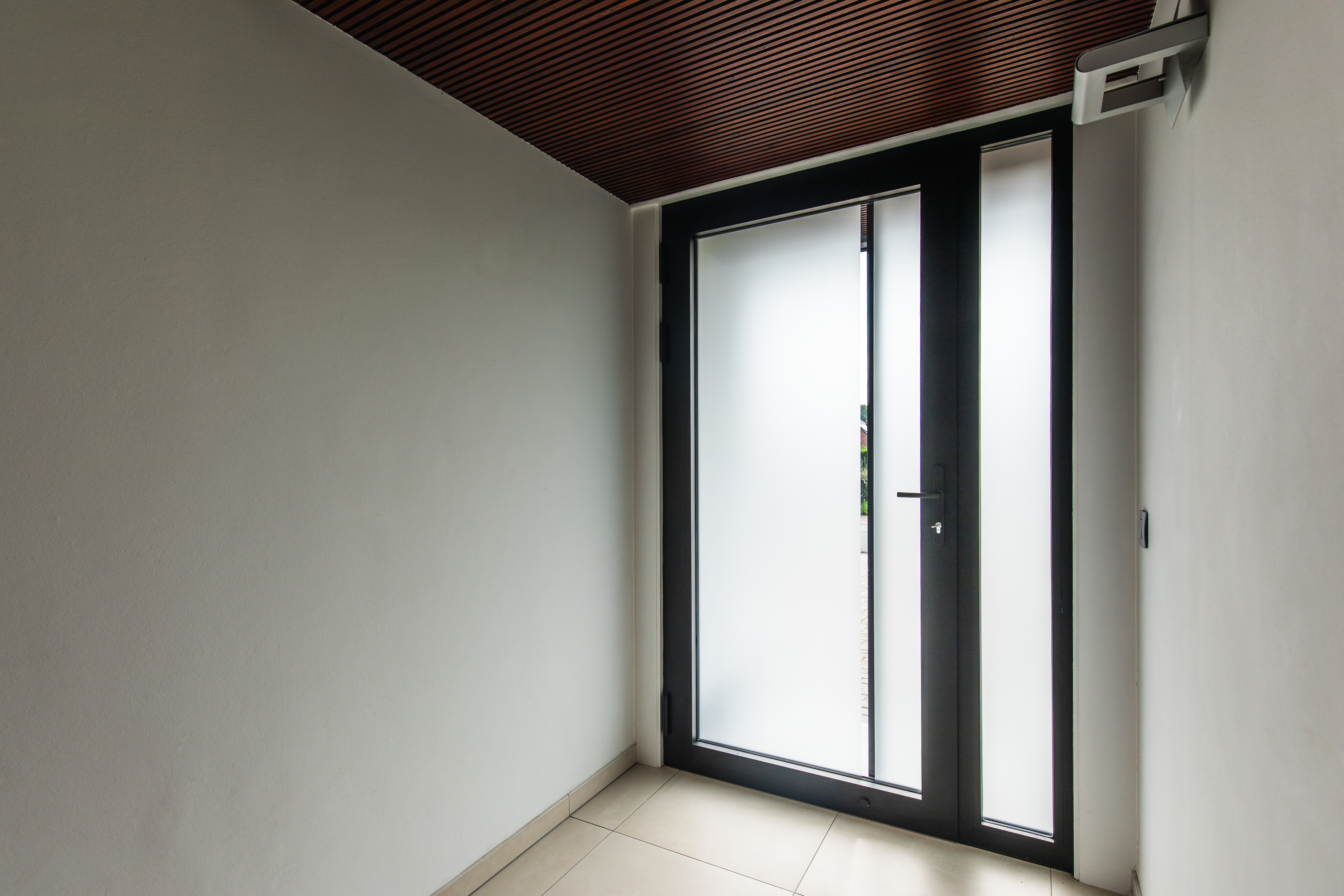 Zwarte aluminium voordeur met beglaasd deurblad en zijlichten in een moderne inkomhal van een gerenoveerde woning.