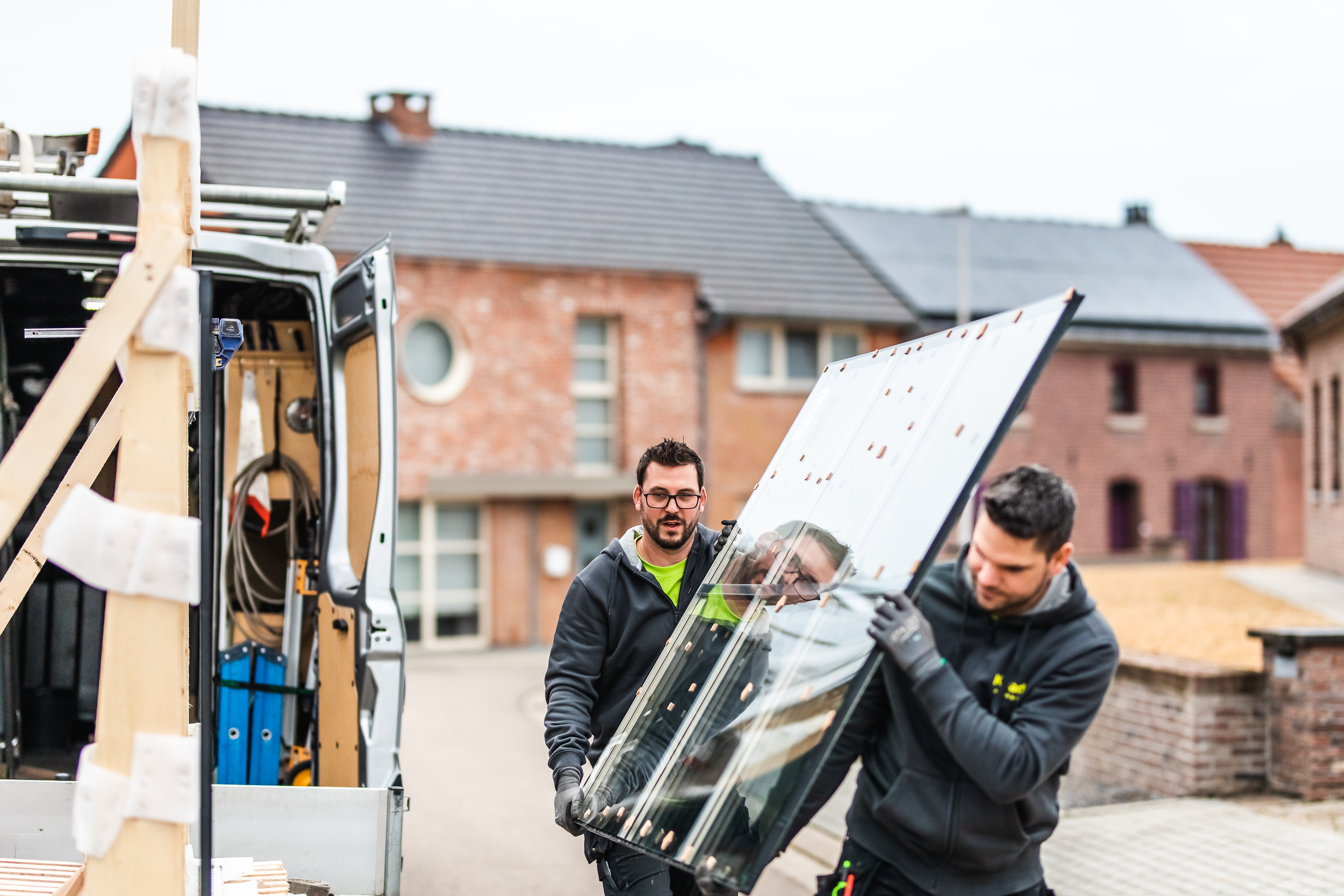 Twee plaatsers dragen een groot raam naar een bestelwagen, met bakstenen rijwoningen op de achtergrond.