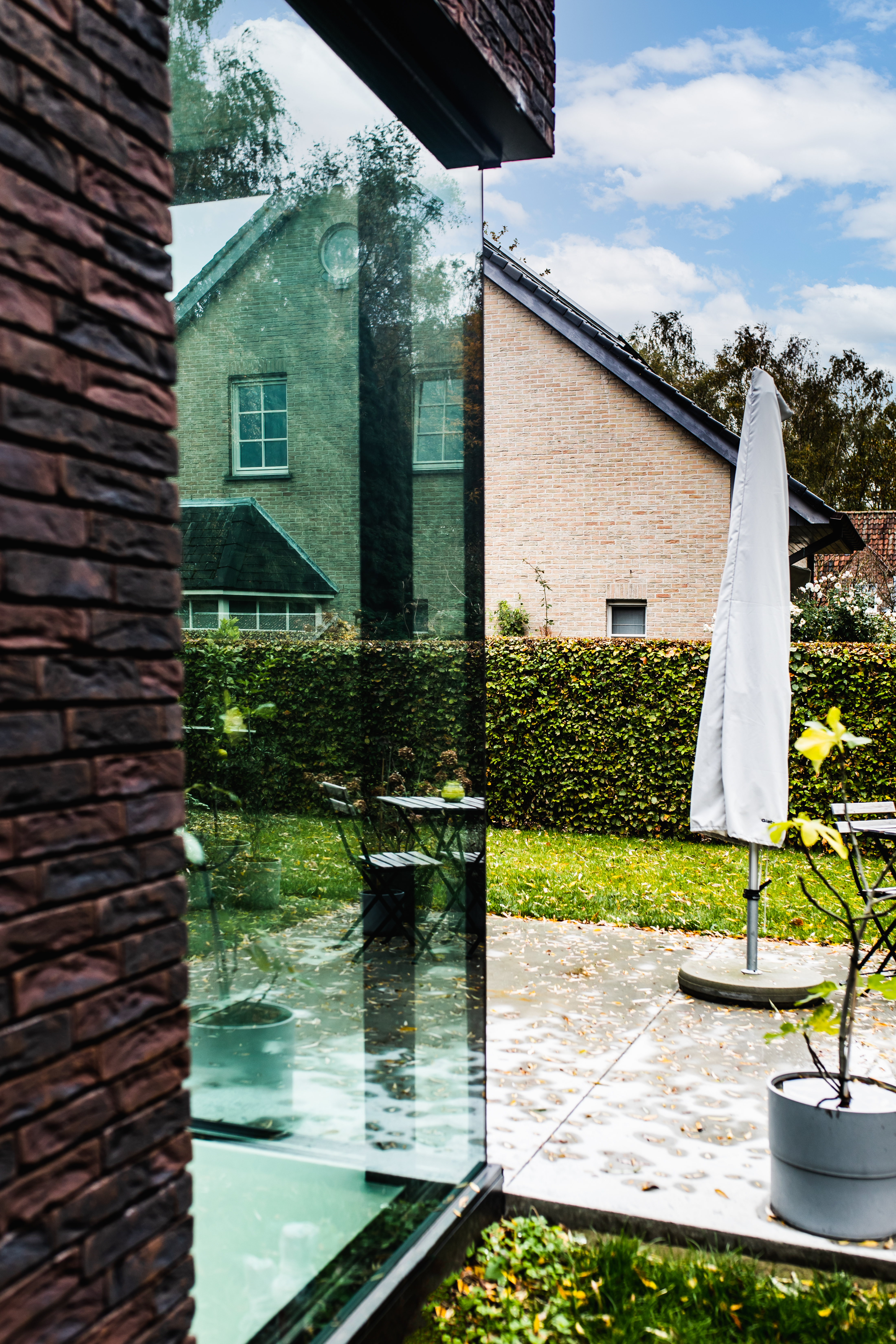 Groot zwart raam in een moderne open bebouwing met bakstenen gevel, terras, tuin en een lichte parasol naast een glazen gevel.