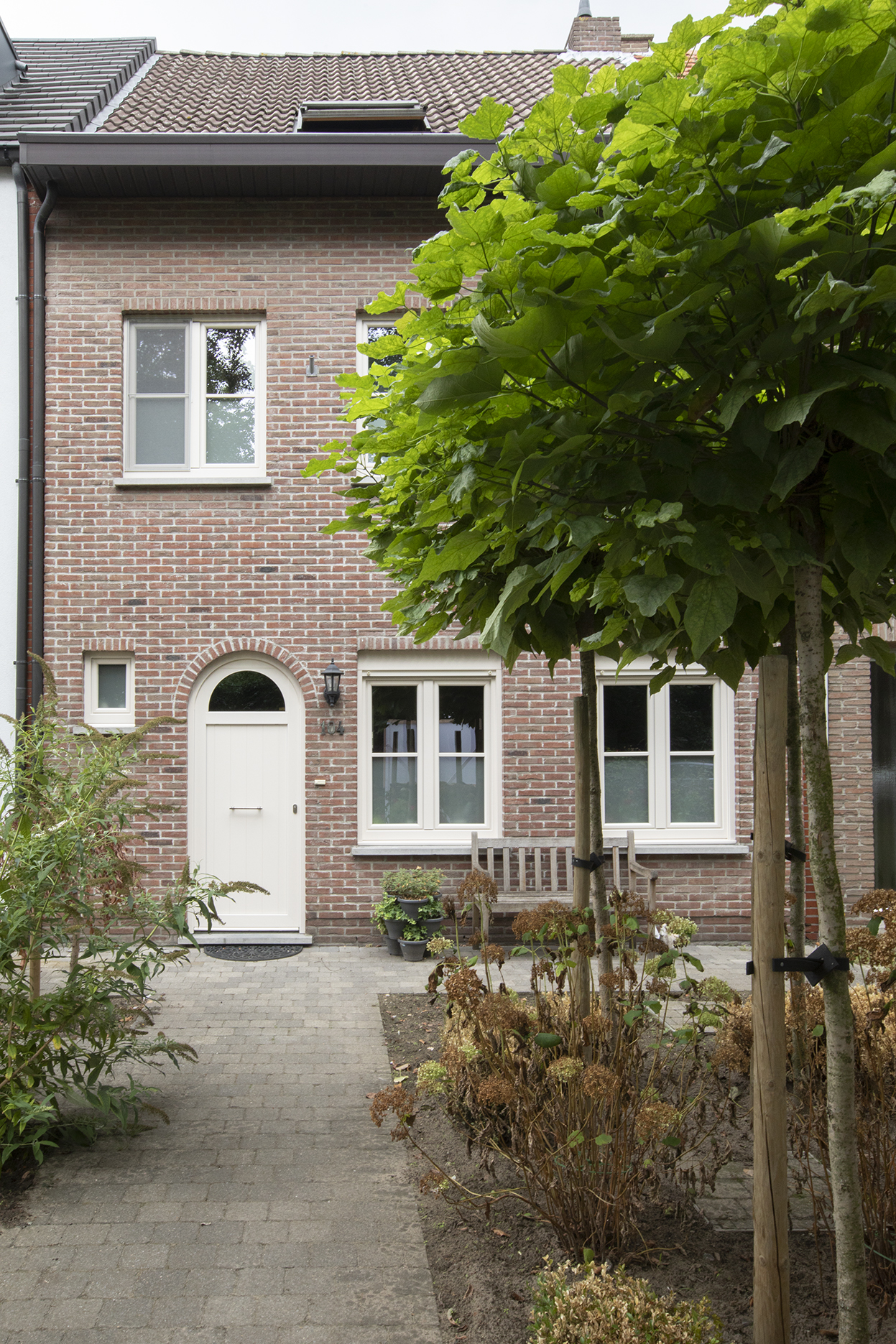 Façade en briques rénovée avec fenêtre double en PVC couleur crème, porte d’entrée avec imposte et volet roulant en applique, dans un jardin avant verdoyant.