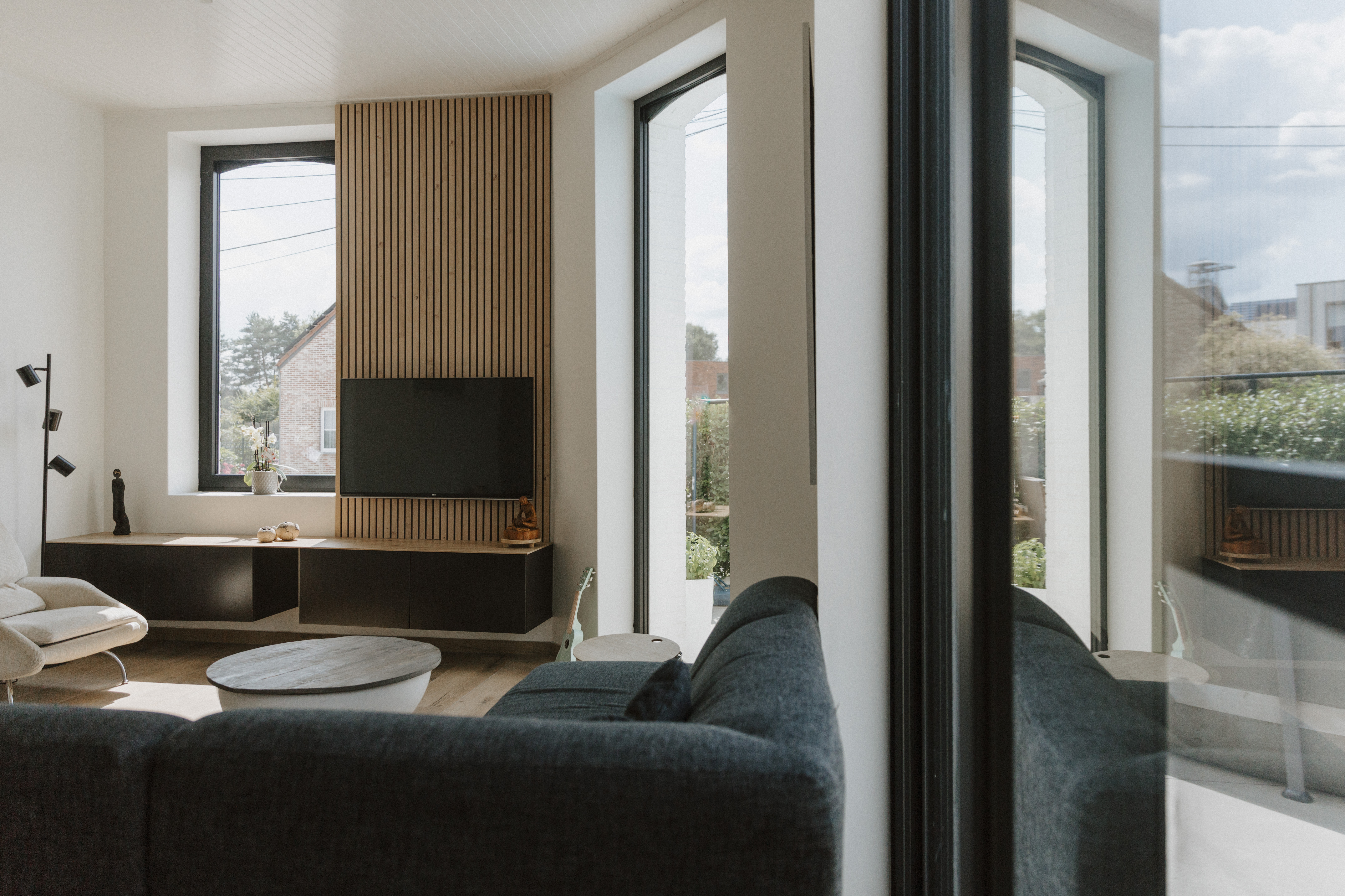 Lichte woonkamer met diepzwarte raamprofielen: een vast raam en een draai-kipraam naast de zithoek en tv-wand met houten latten.
