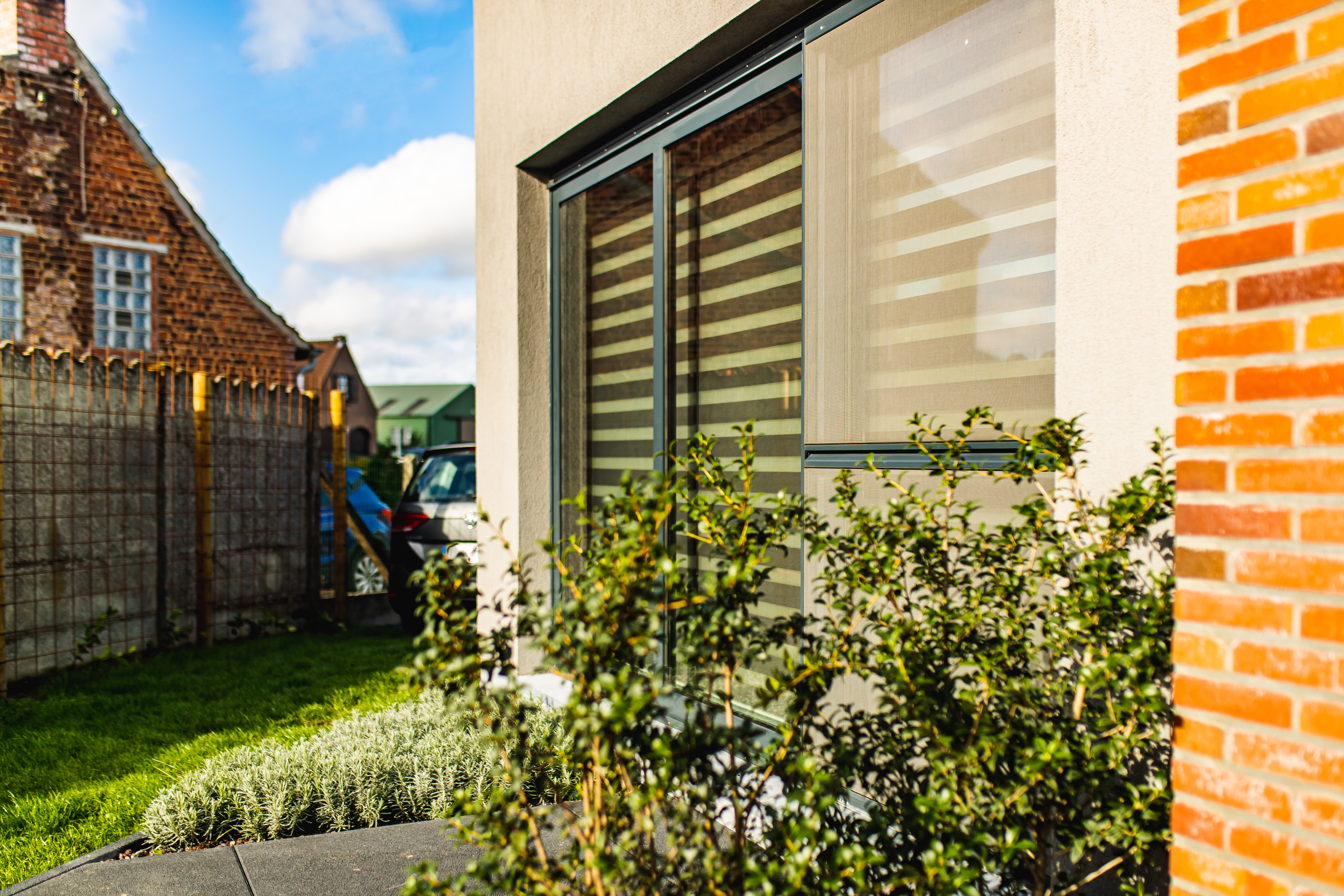 Antracietgrijze raamprofielen met grote ramen in een nieuwbouwwoning, met een gemetselde gevel en groene voortuin naast een open bebouwing.