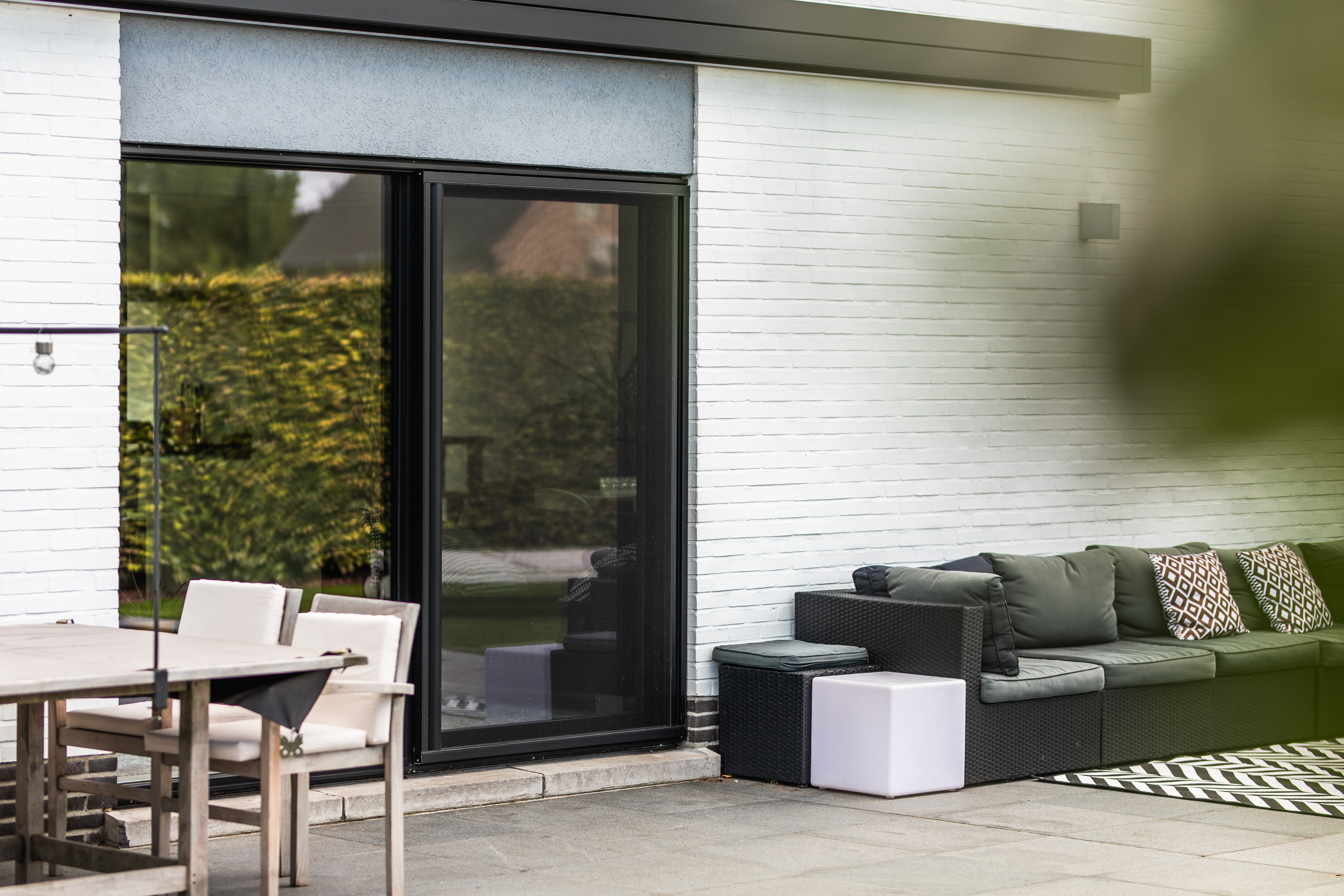 Zwart aluminium schuifvliegenraam van het ALU Visoglide Monorail-systeem op het terras van een moderne open bebouwing, naast een zithoek en eettafel.