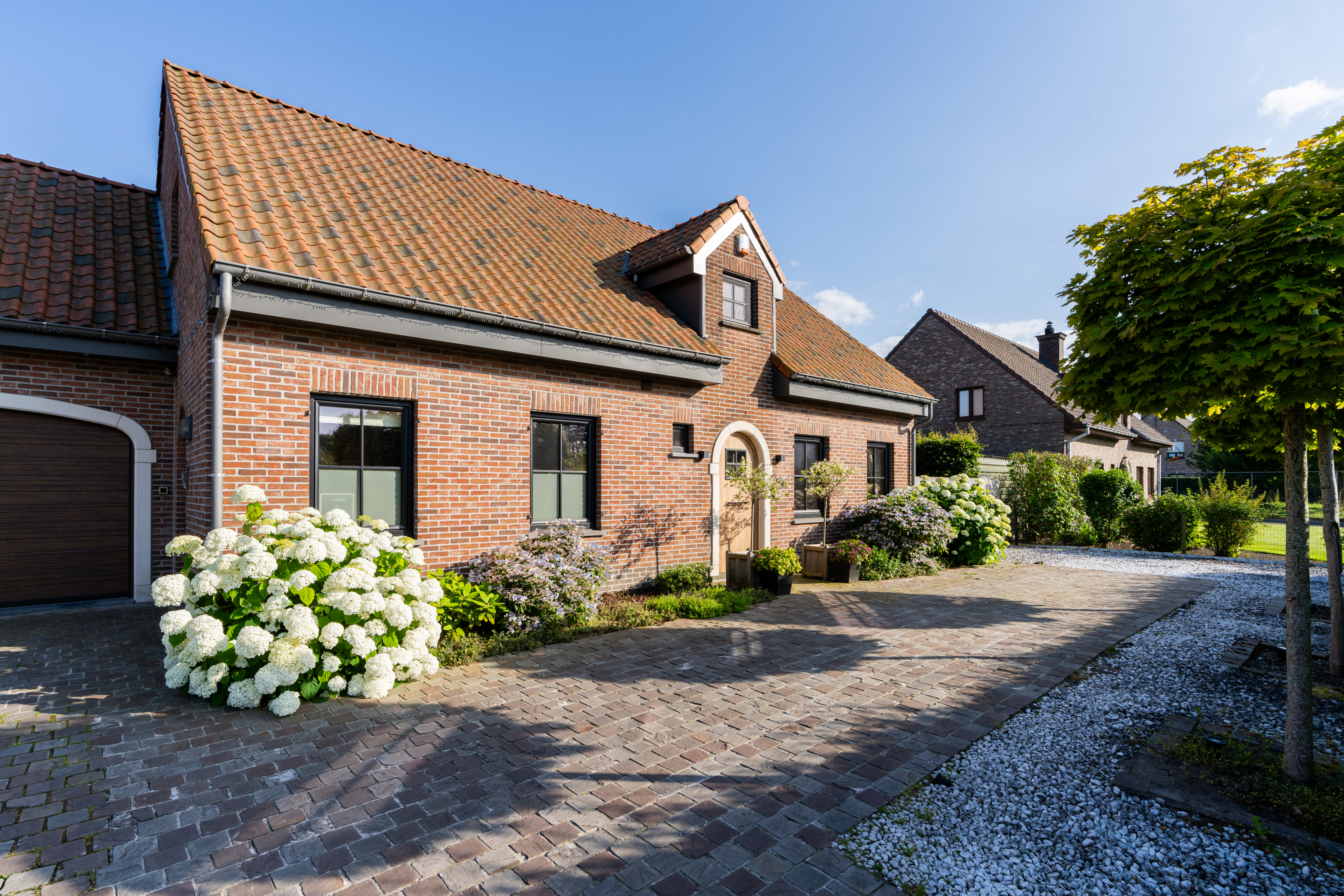 Bakstenen gevel van een charmante woning met pannendak, ramen en een gebogen inkom, omgeven door hortensia’s, een gekasseide oprit en een verzorgde tuin.