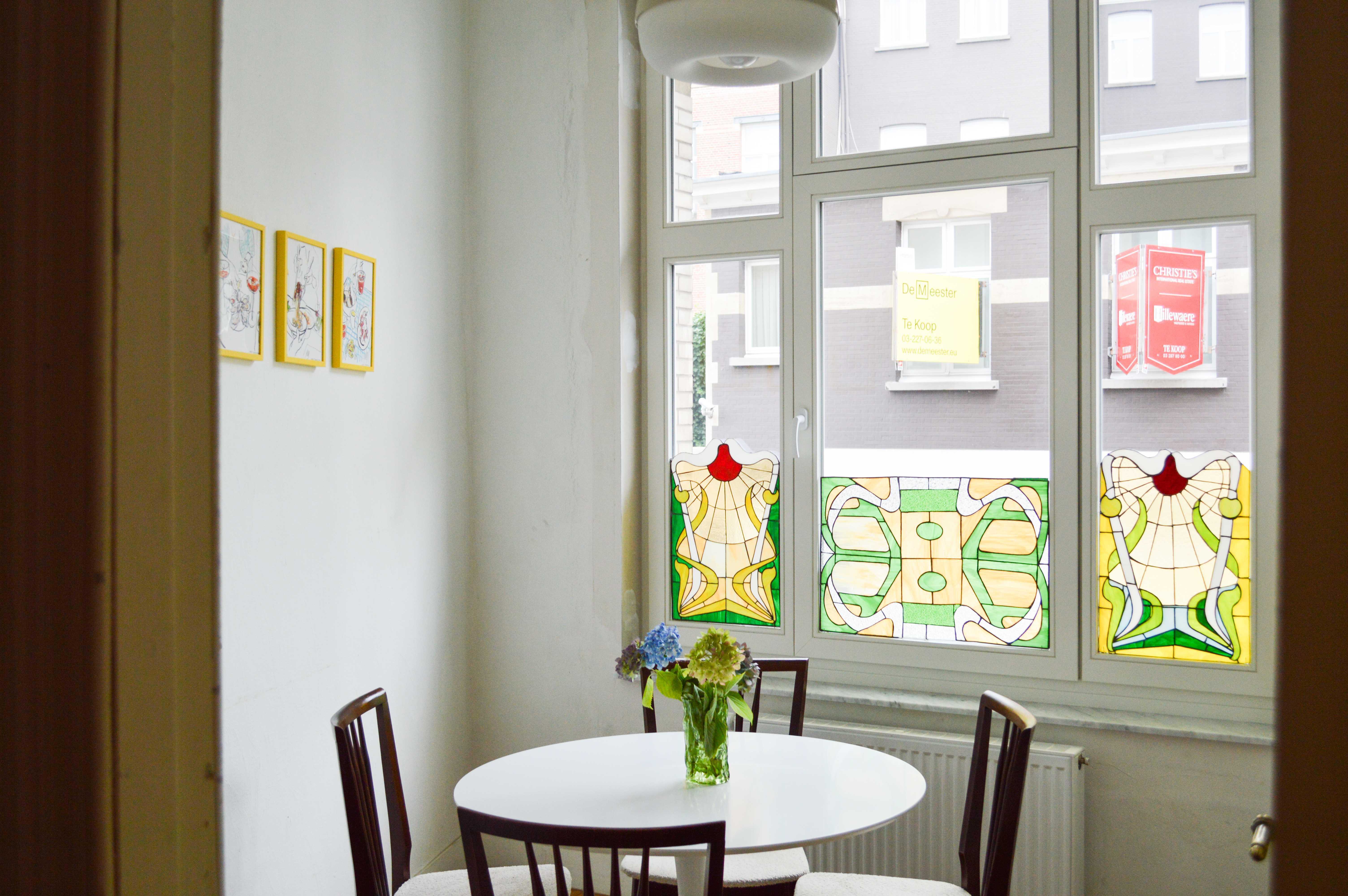 Gerenoveerd interieur met verfijnd klassiek, wit afgelakt raamensemble met een vast raam, draai-kipraam en bovenlicht, met kleurrijke glas-in-loodpanelen uitkijkend op gesloten bebouwing.