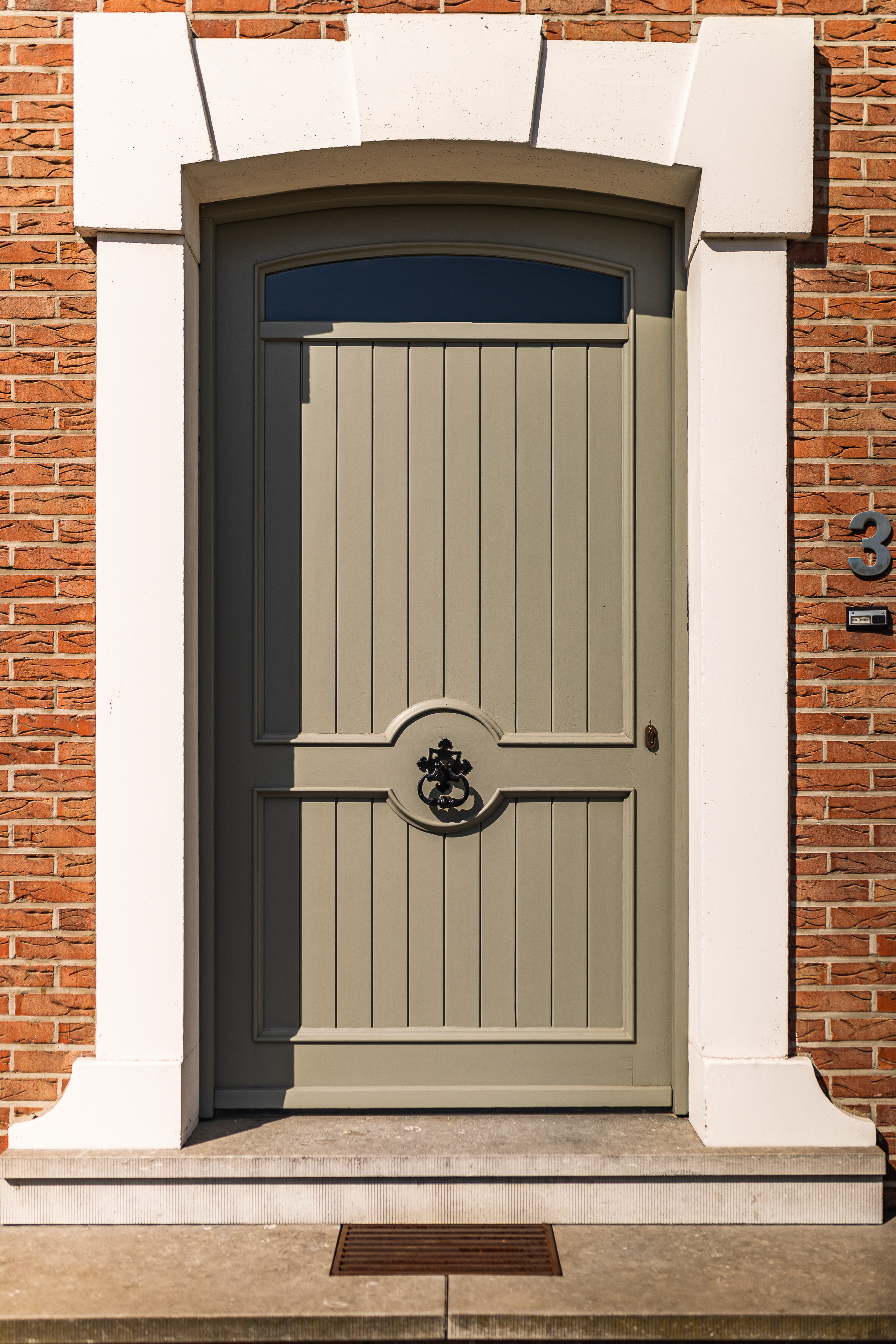Porte d’entrée intemporelle en PVC gris chaud, avec panneau et imposte vitrée, encadrée d’éléments blancs dans une façade en briques rénovée.