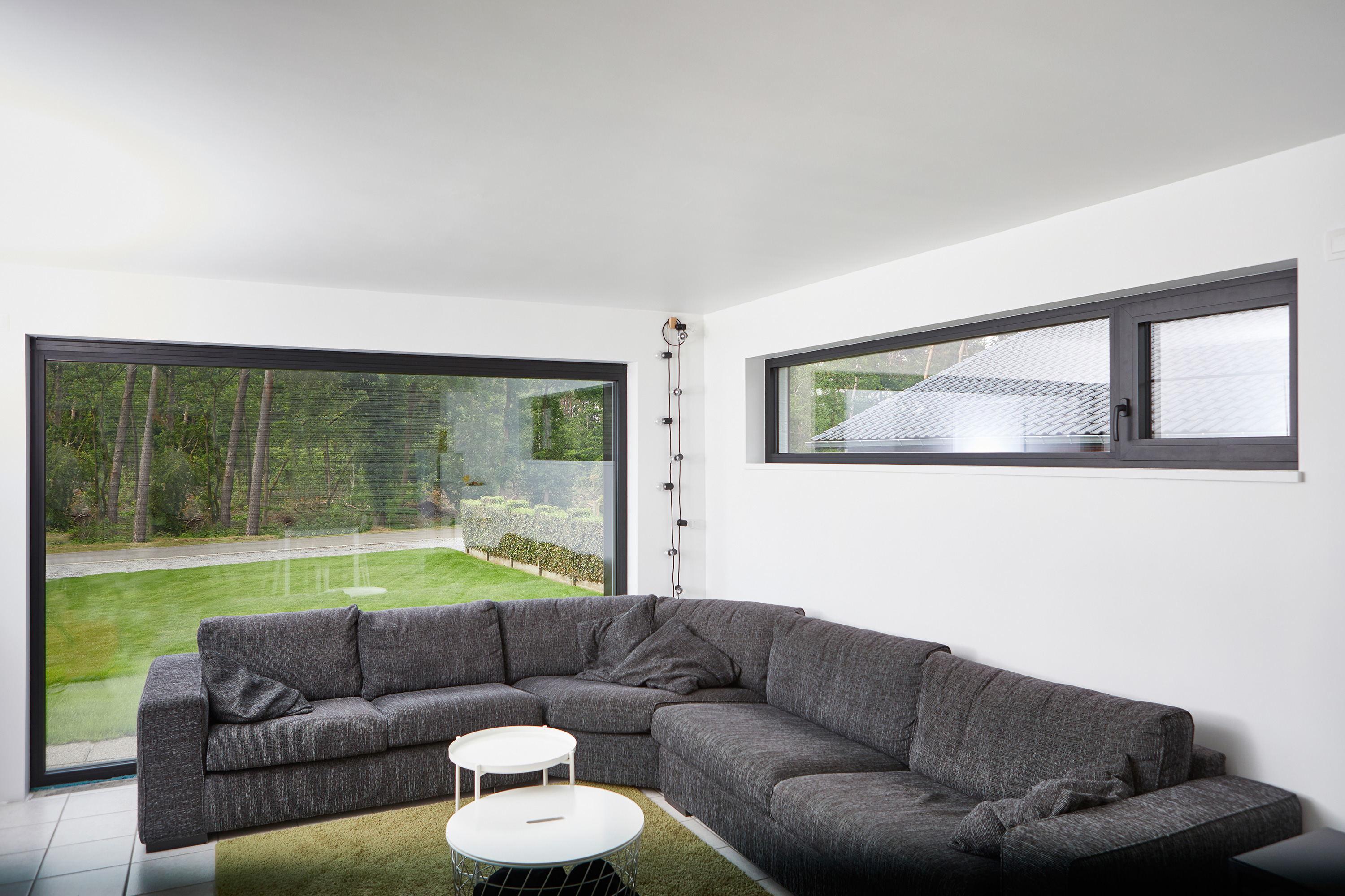 Modern gerenoveerde leefruimte met grote zwarte vaste ramen in steellook, met zicht op de groene tuin en een ruime hoekzetel.
