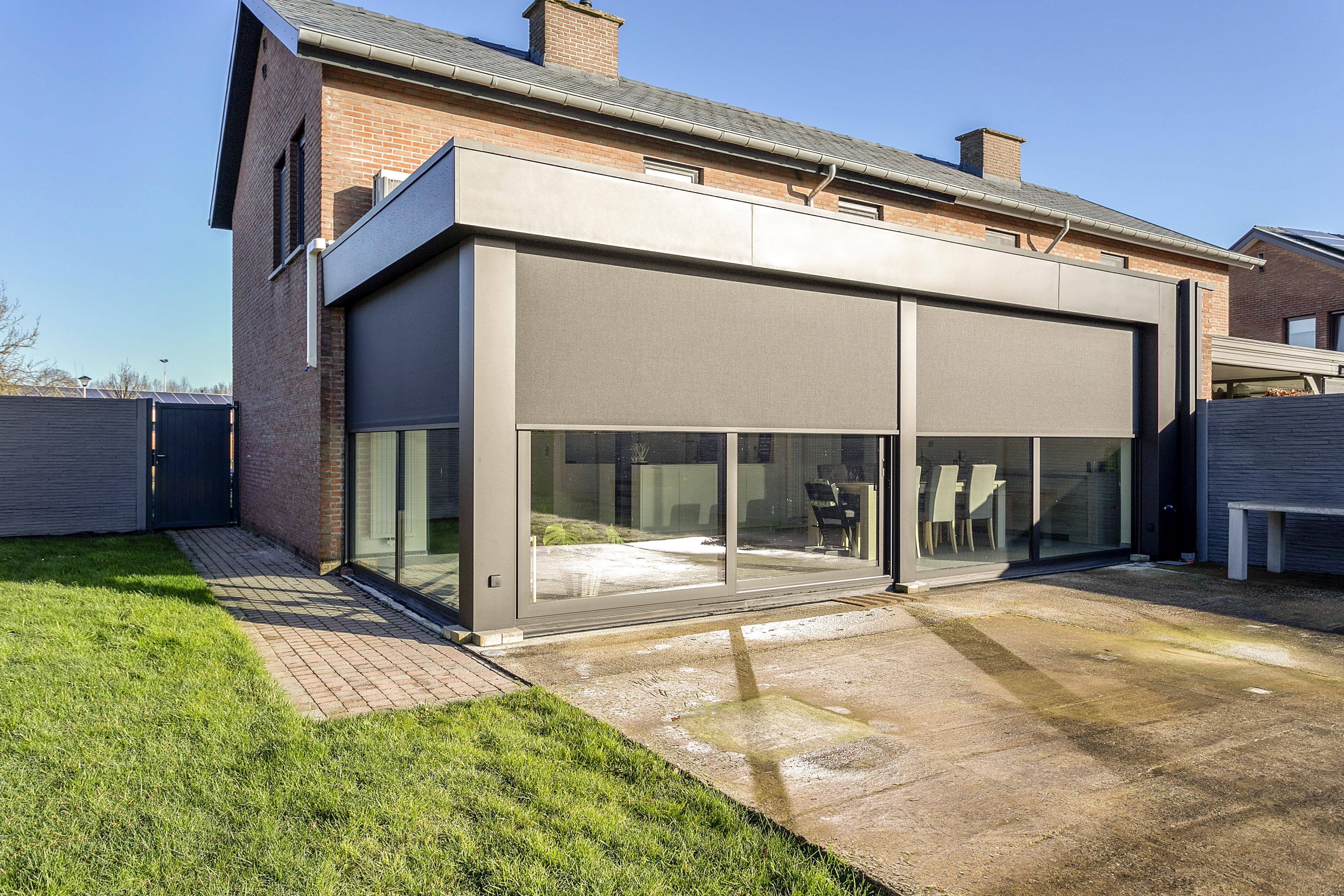 Groot vast raam met ingebouwde screens en zwarte aluminiumprofielen aan de gerenoveerde halfopen woning, naast een gedeeltelijk beglaasde aanbouw.