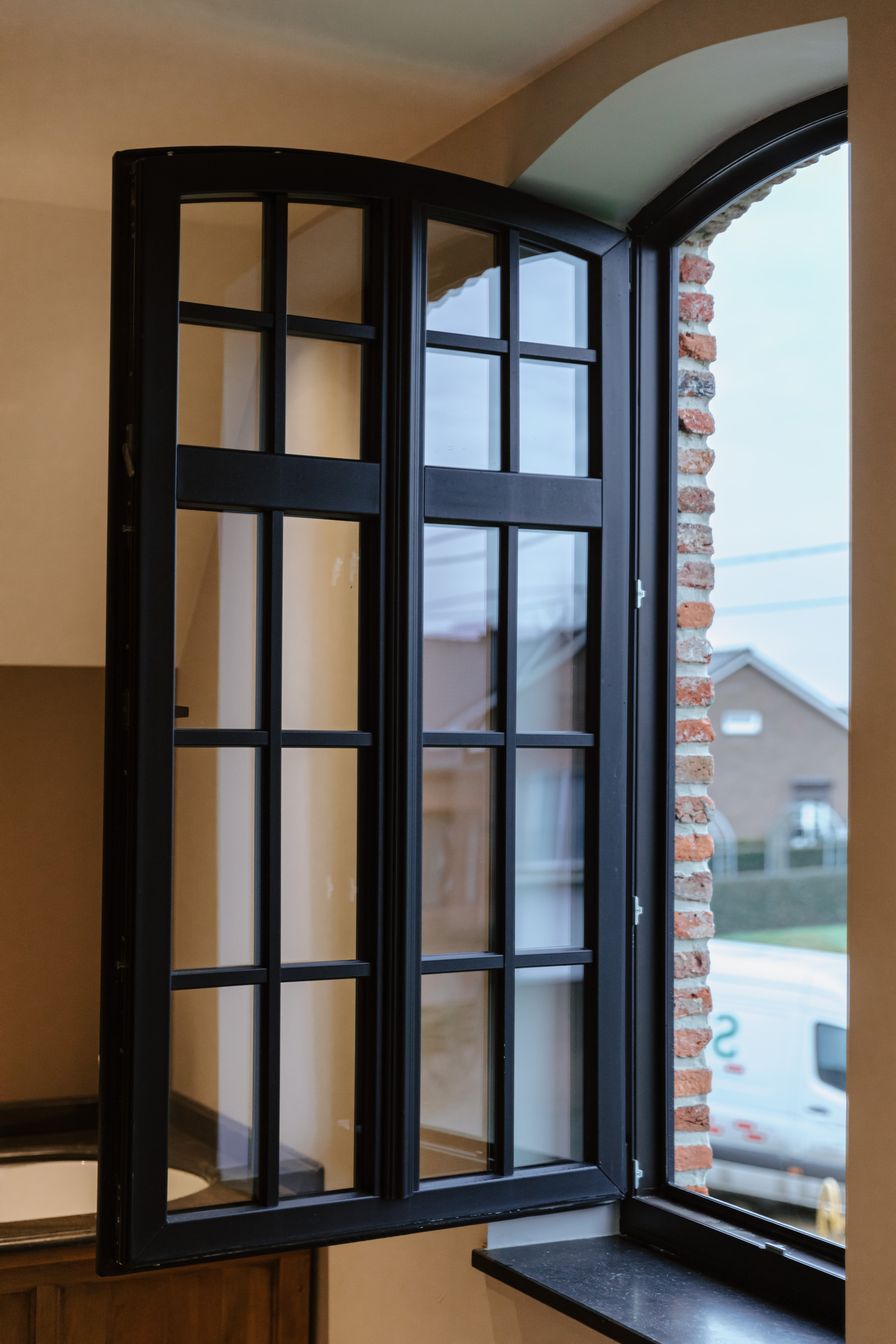 Mat zwart draai-kipraam met klassieke roedeverdeling, geopend in een warm interieur; door de ronde boog zijn baksteen en een woonwijk zichtbaar.