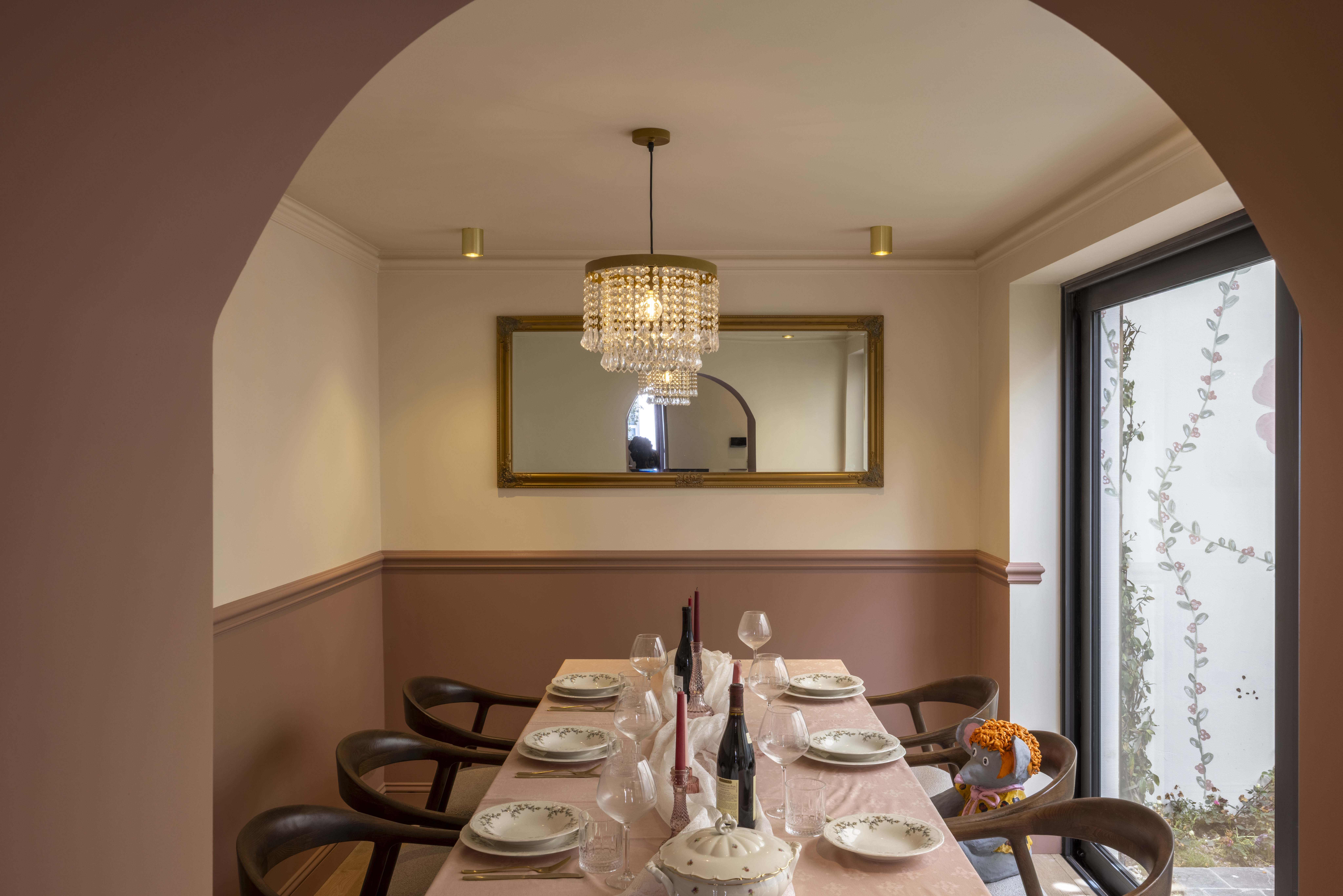 Eetkamer met gedekte houten tafel, spiegel en kristallen hanglamp, naast een bronzen aluminium schuifraam met decoratieve beglazing.