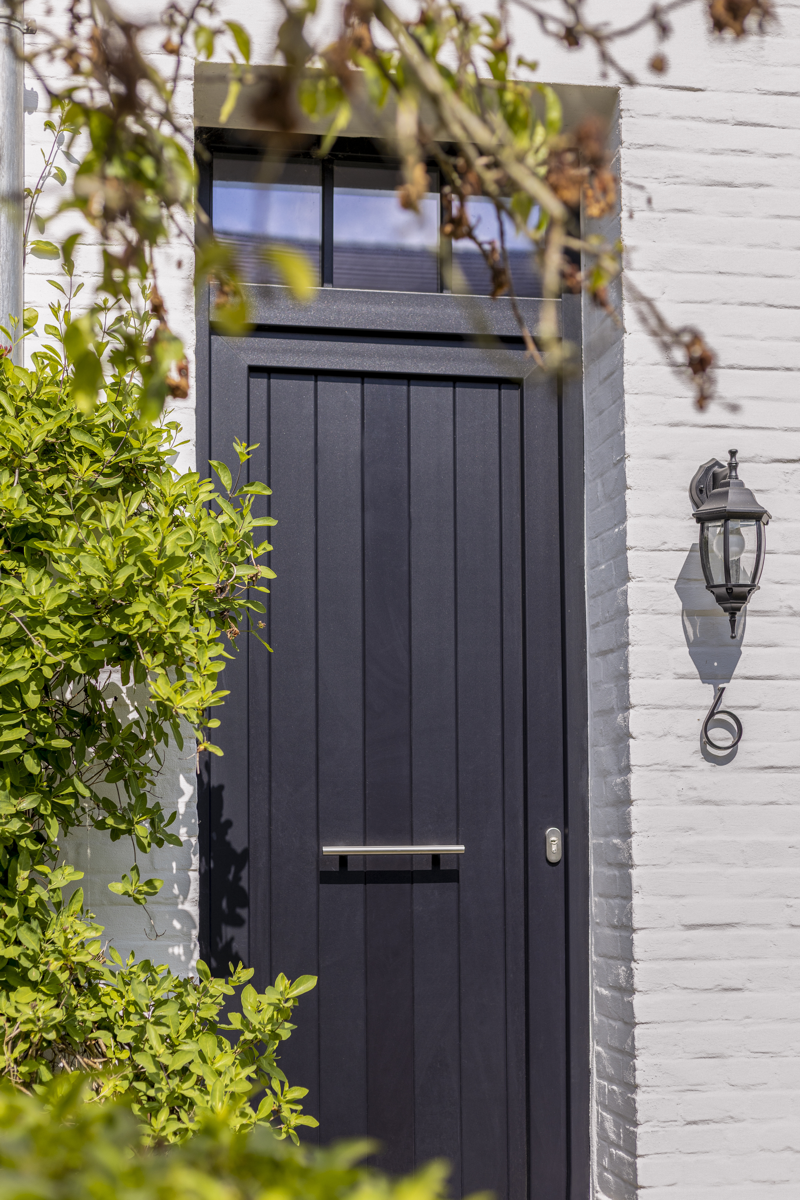 Porte d’entrée à panneaux en PVC Impakt PLUS noir mat, avec imposte et poignée horizontale, dans la façade en briques blanches d’une maison jumelée rénovée.