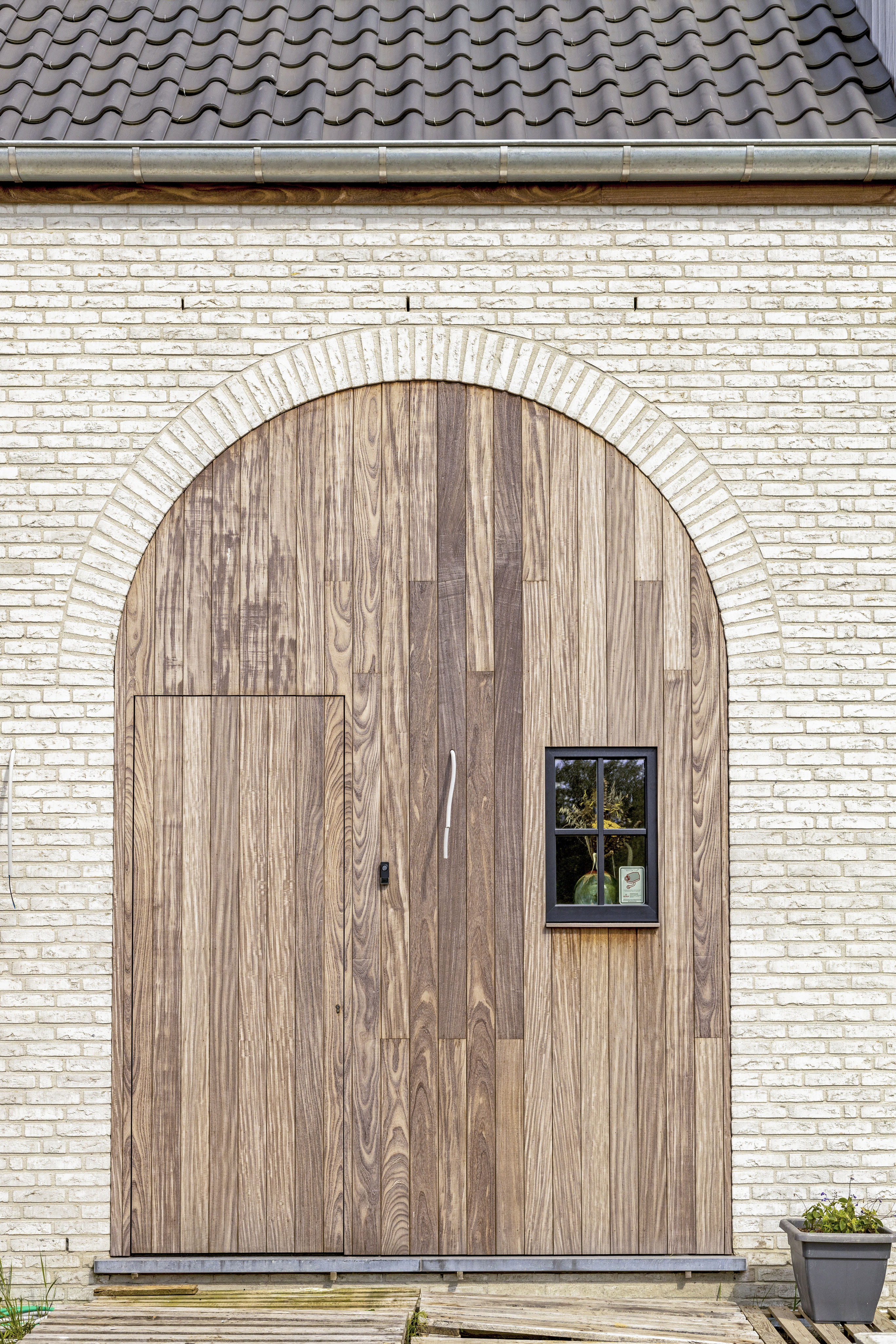 Gevel van een nieuwbouwwoning in warme landelijke stijl, met lichte bakstenen, een gewelfde houten deur, klein matzwart raam en pannendak.