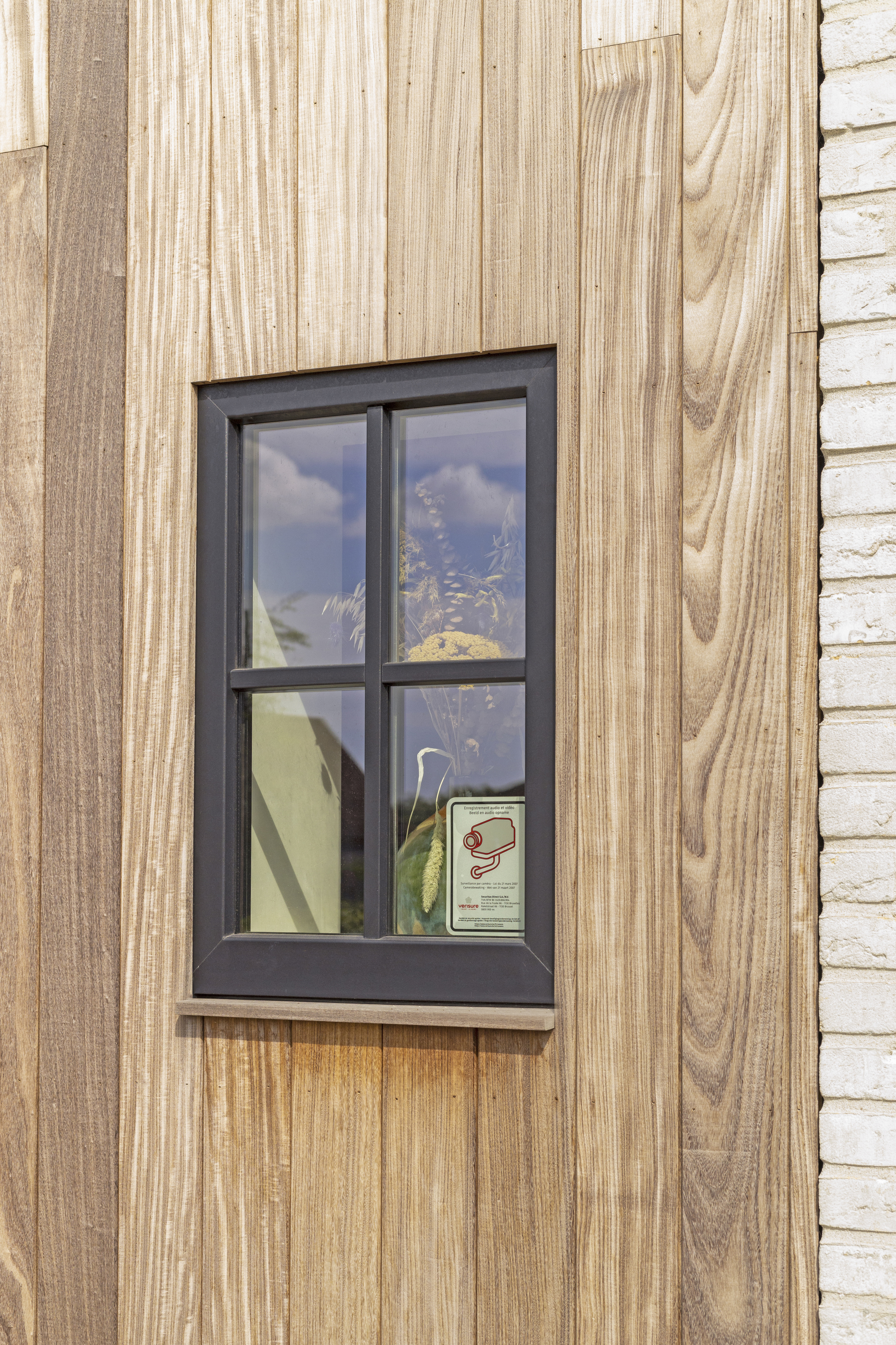 Smal, matzwart raam in een warme houten gevel van een landelijke nieuwbouw, naast een lichte bakstenen muur.