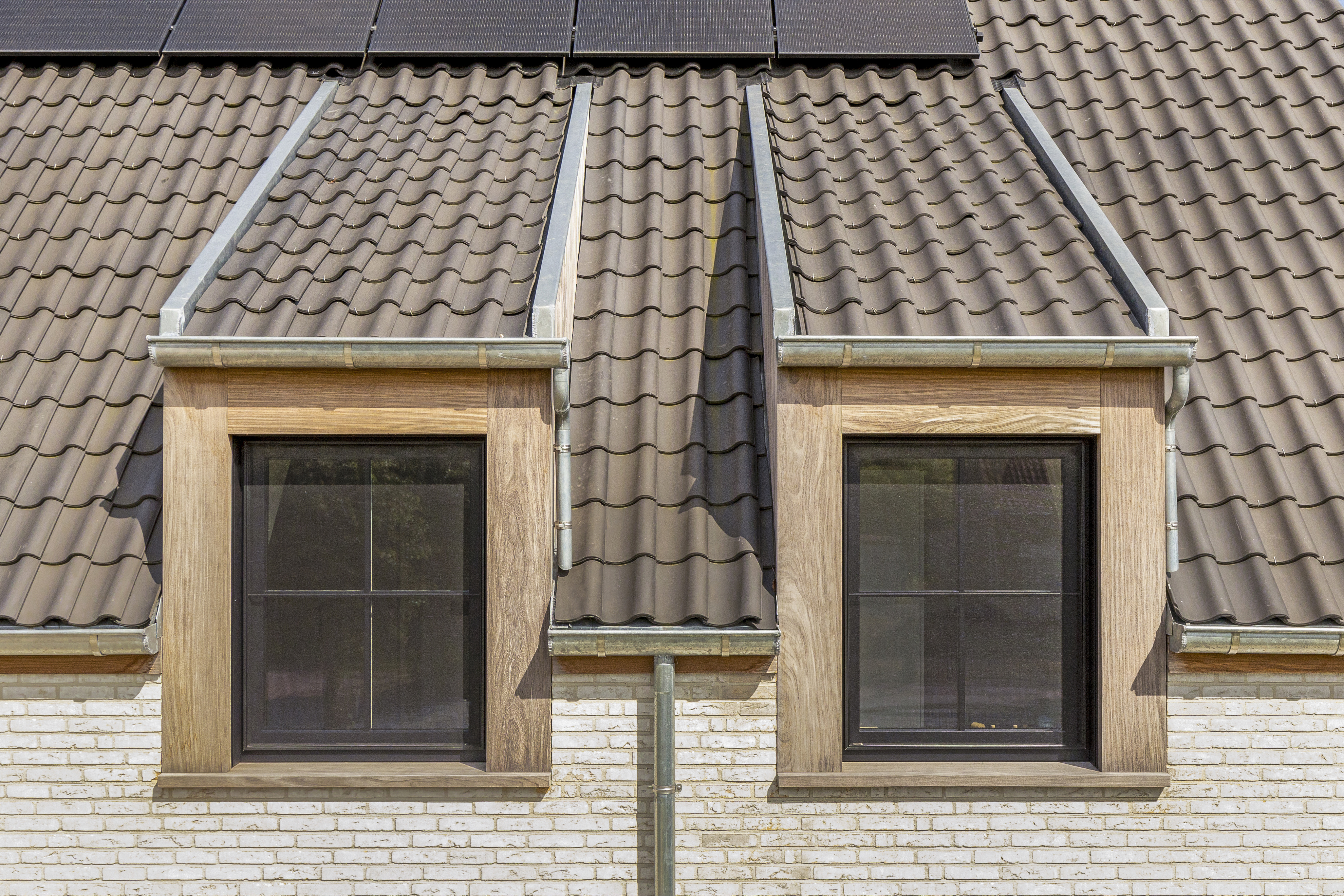 Twee matzwarte draai-kipramen met vliegenramen in houten dakkapellen van een warm landelijke woning, boven een lichte bakstenen gevel.