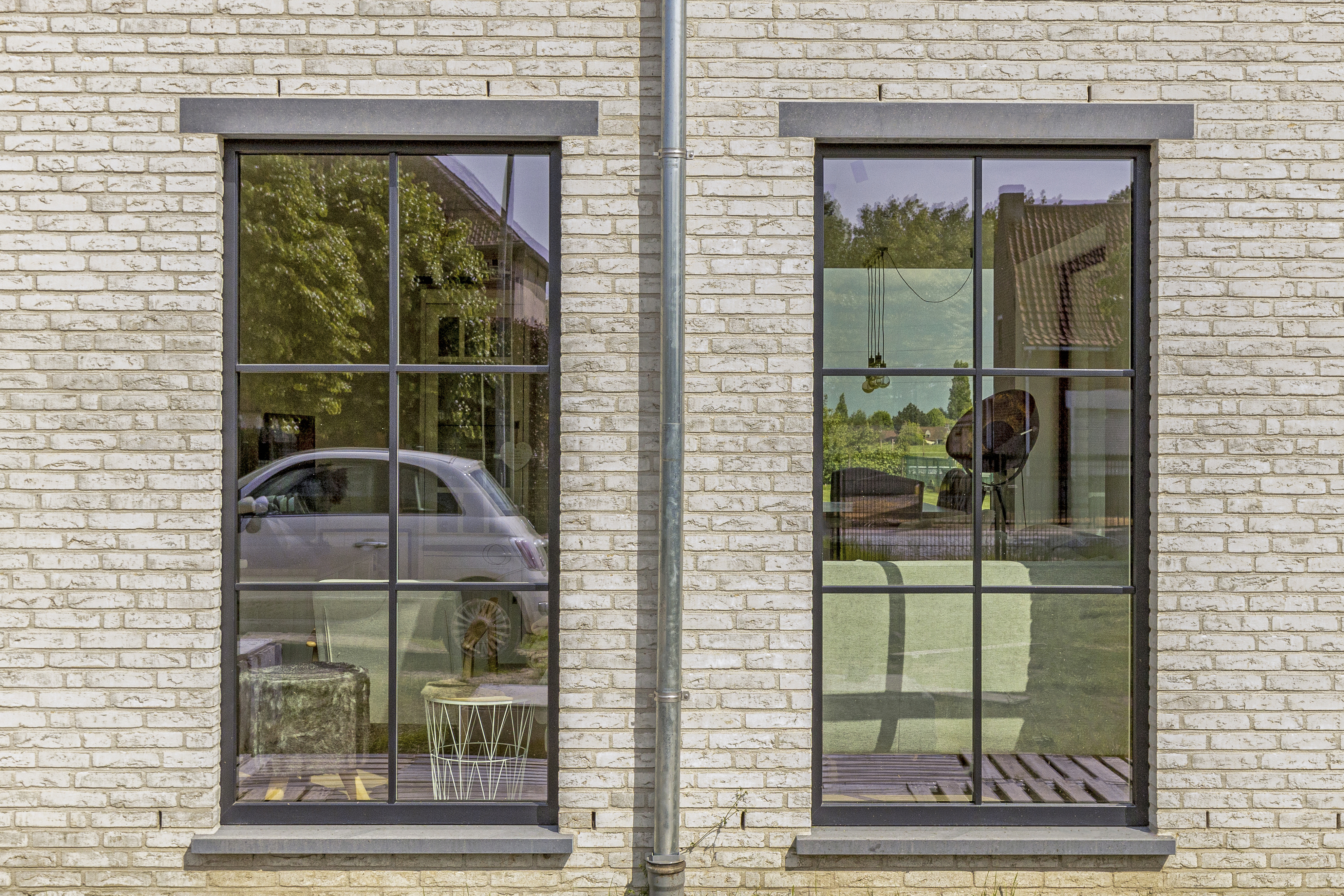 Twee hoge, matzwarte vaste ramen in een lichte bakstenen gevel van een warm landelijk vormgegeven nieuwbouwwoning, met fijne raamverdelingen.