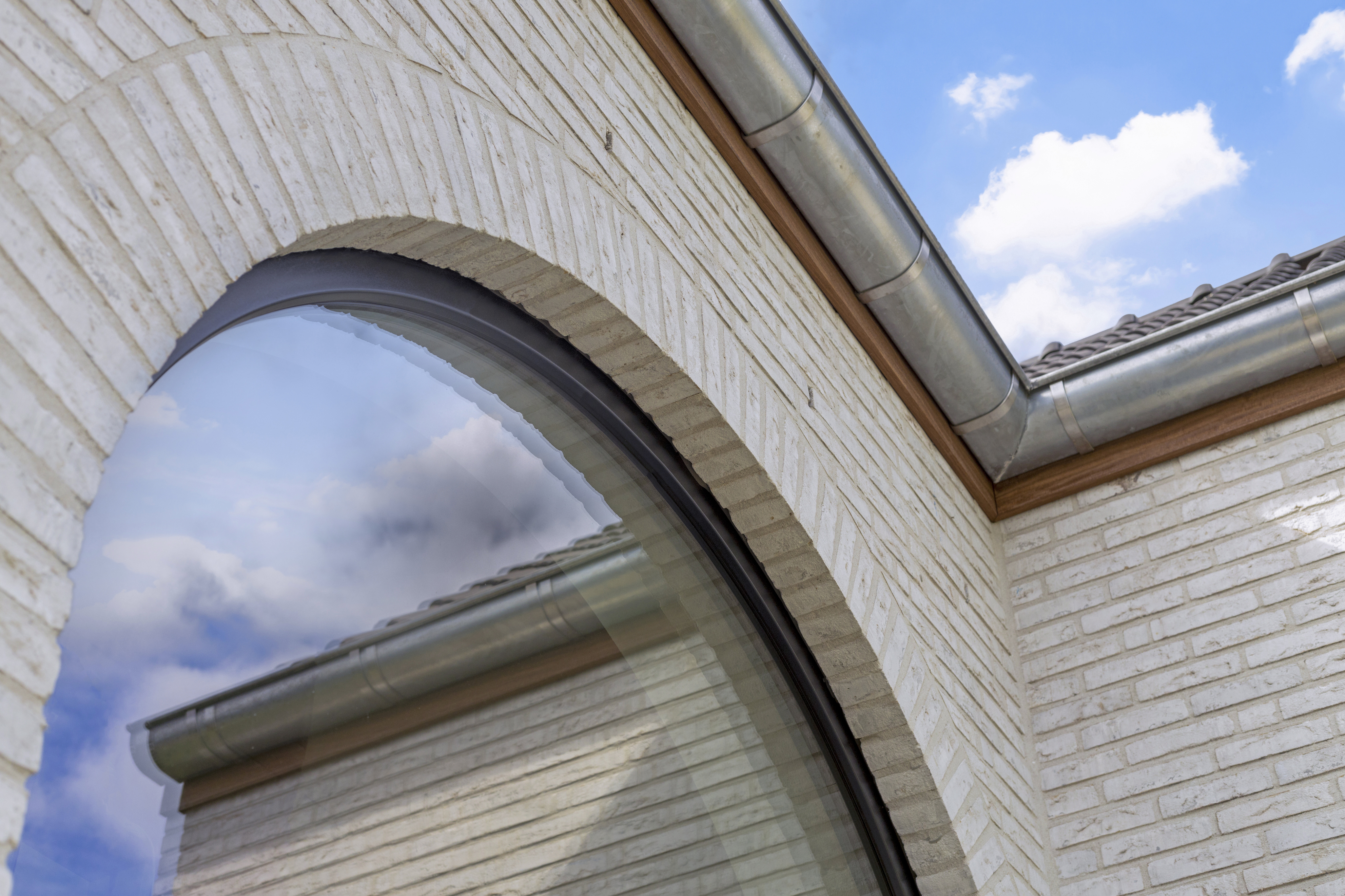 Gebogen vast raam met matzwart raamprofiel in een witte bakstenen gevel van een landelijke nieuwbouwwoning; de beglazing weerspiegelt de dakgoot, bakstenen en lucht.