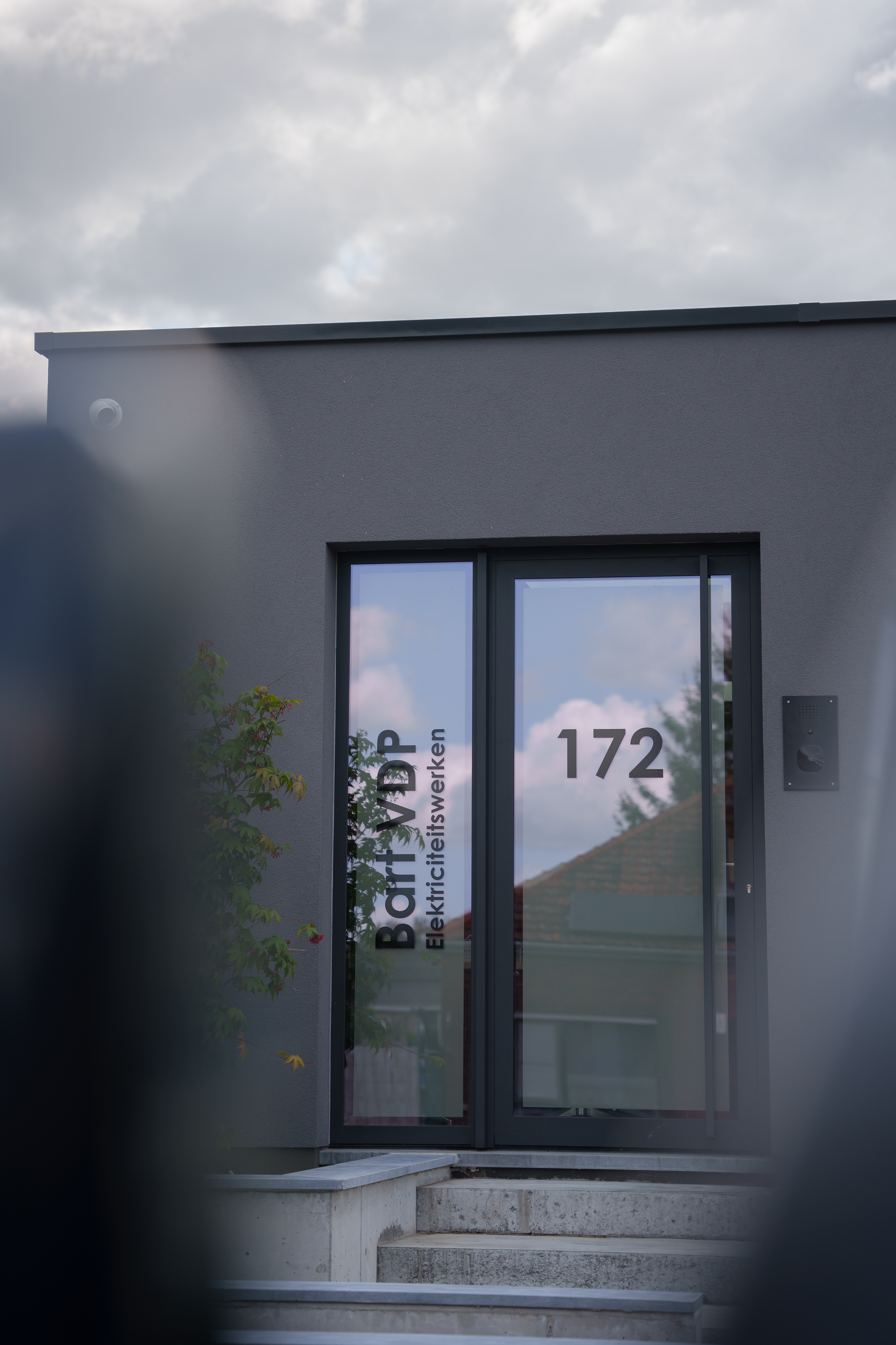 Moderne nieuwbouwwoning met een diepzwarte aluminium beglaasde deur en zijlicht in een donkere gevel, bereikbaar via een buitentrap.
