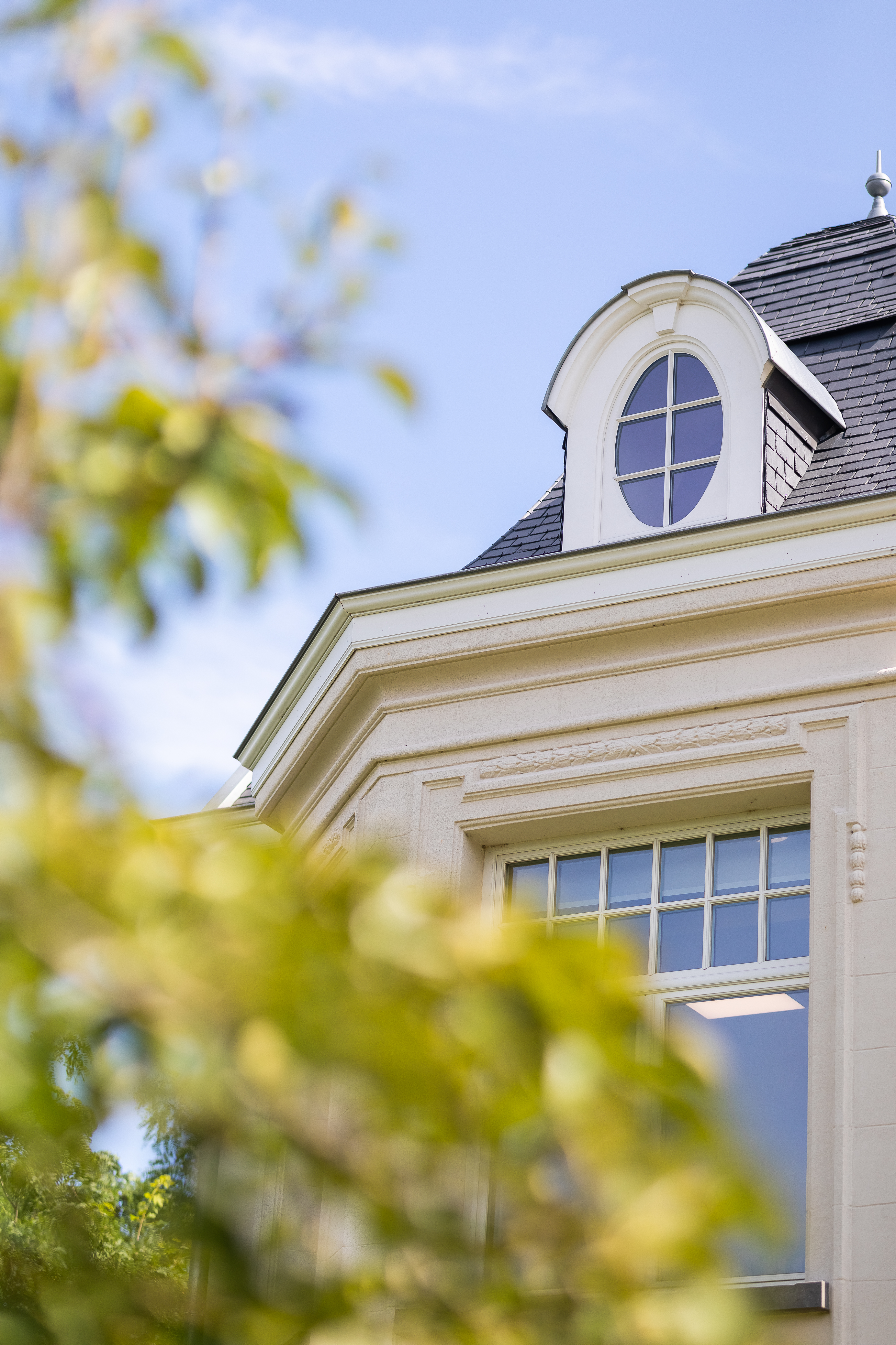 Verfijnd klassieke gevel van een vrijstaande woning in zuiver wit, met een groot raam met roedeverdeling, een rondboograam in de dakopbouw en een donker leien dak, deels omringd door bladeren.