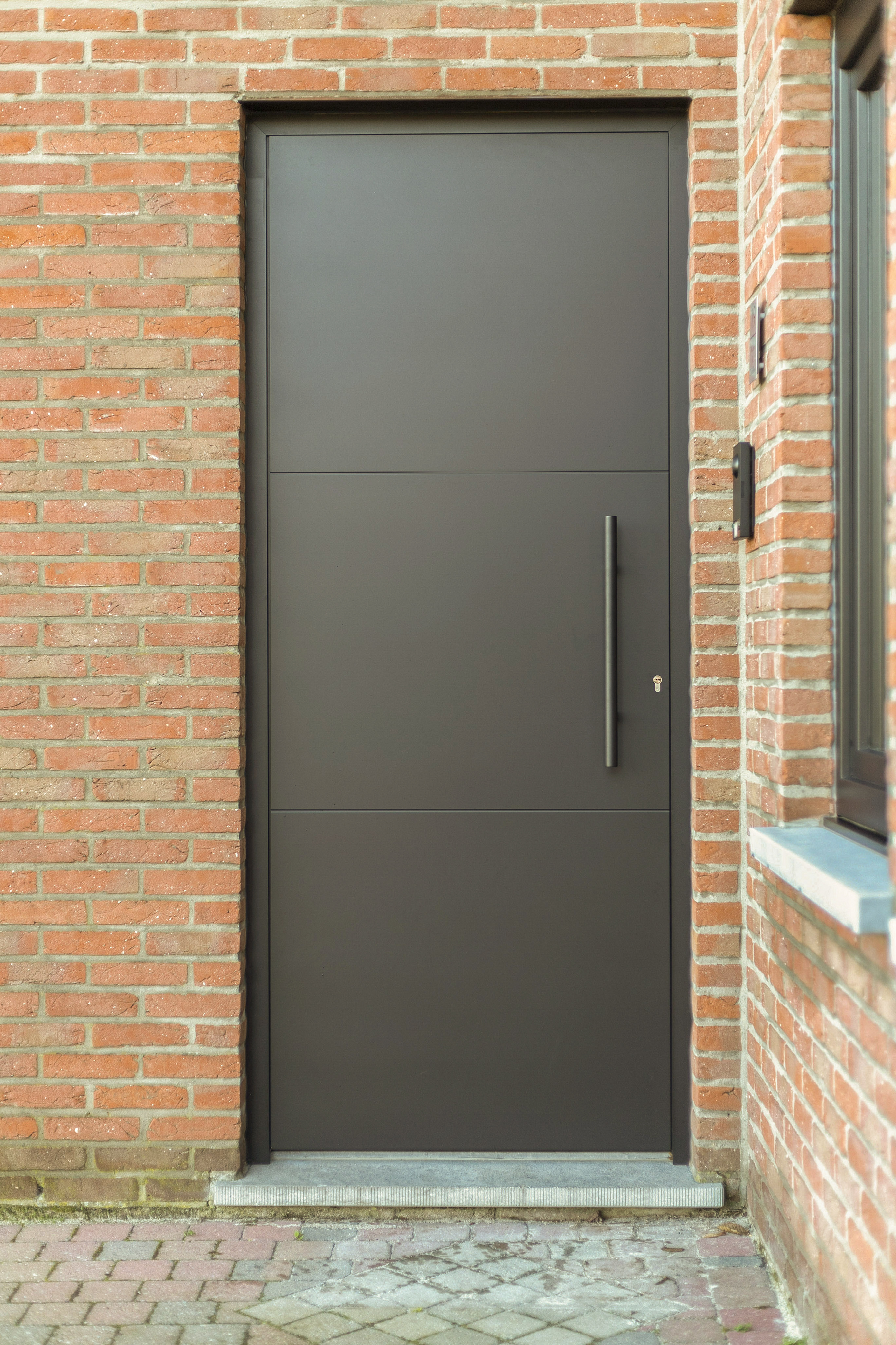 Matzwarte PVC-paneeldeur met lange handgreep in een gerenoveerde gevel van rode baksteen, bij een halfopen bebouwing.