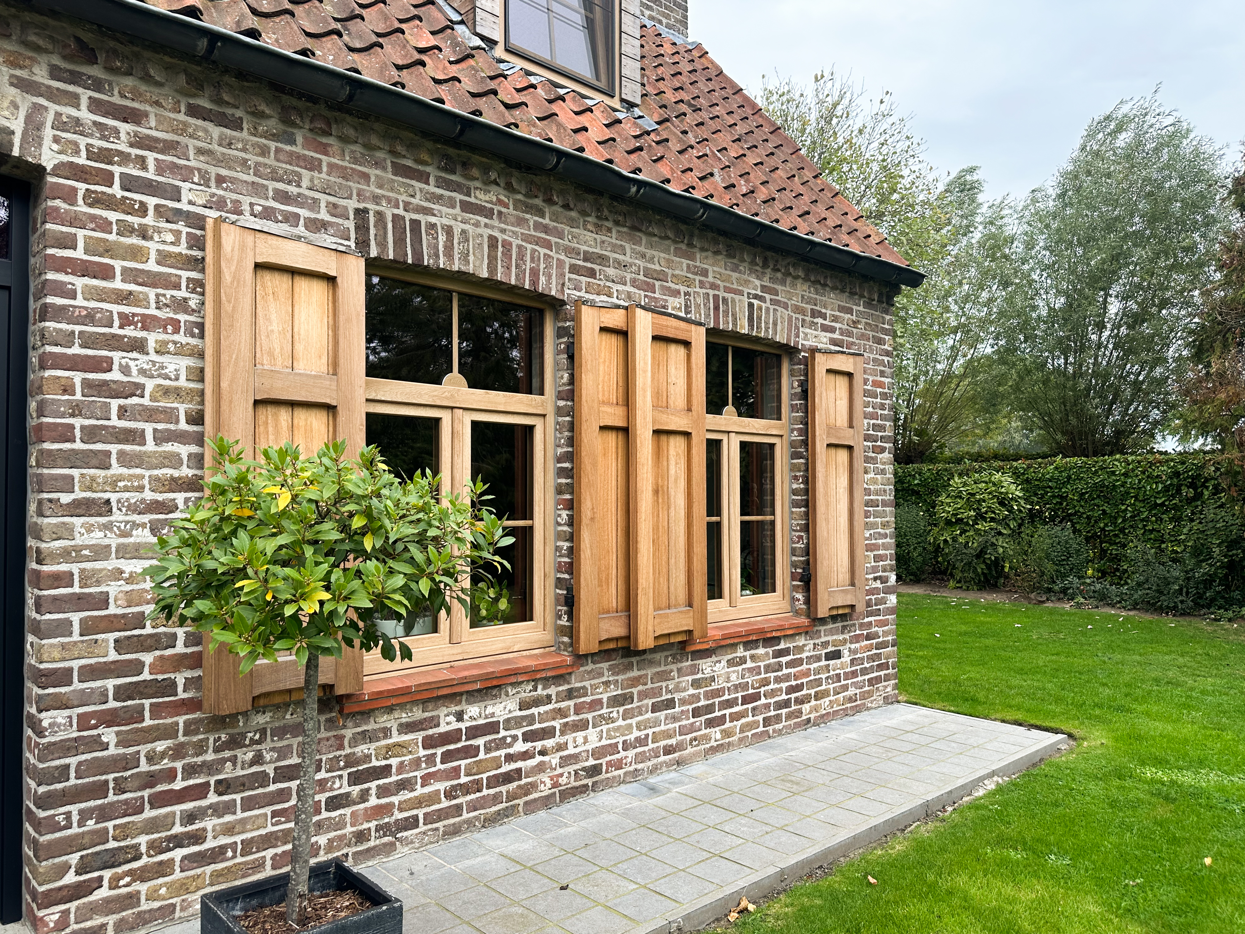 Bakstenen gevel met twee houtkleurige dubbele draai-kiepramen met bovenlichten, rechte profielen en luiken met natuurlijke eiklook, naast een betegeld tuinpad.