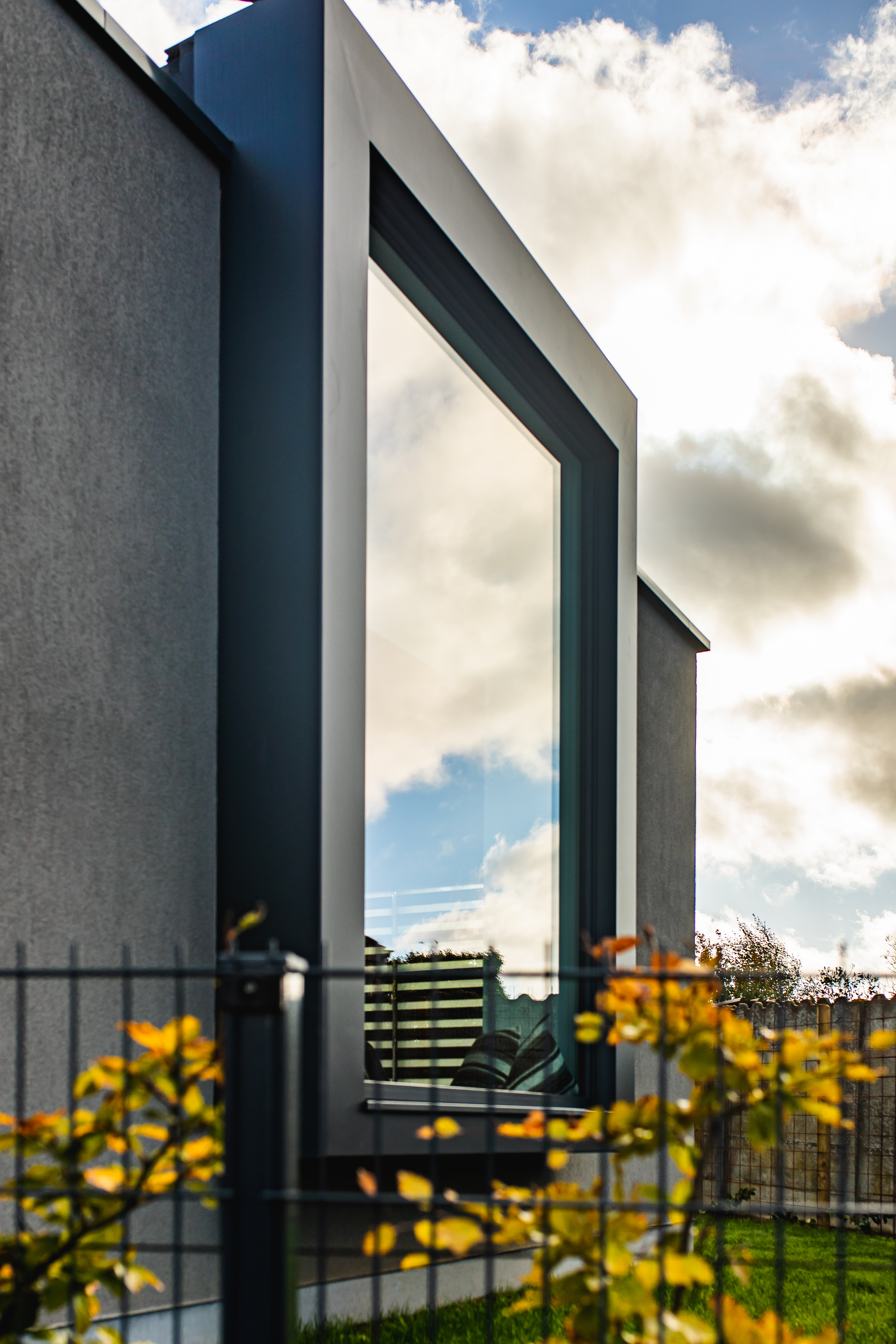 Antracietgrijze gevel van een moderne nieuwbouwwoning met een hoog, smal raam, een zwarte afsluiting en tuinplanten.