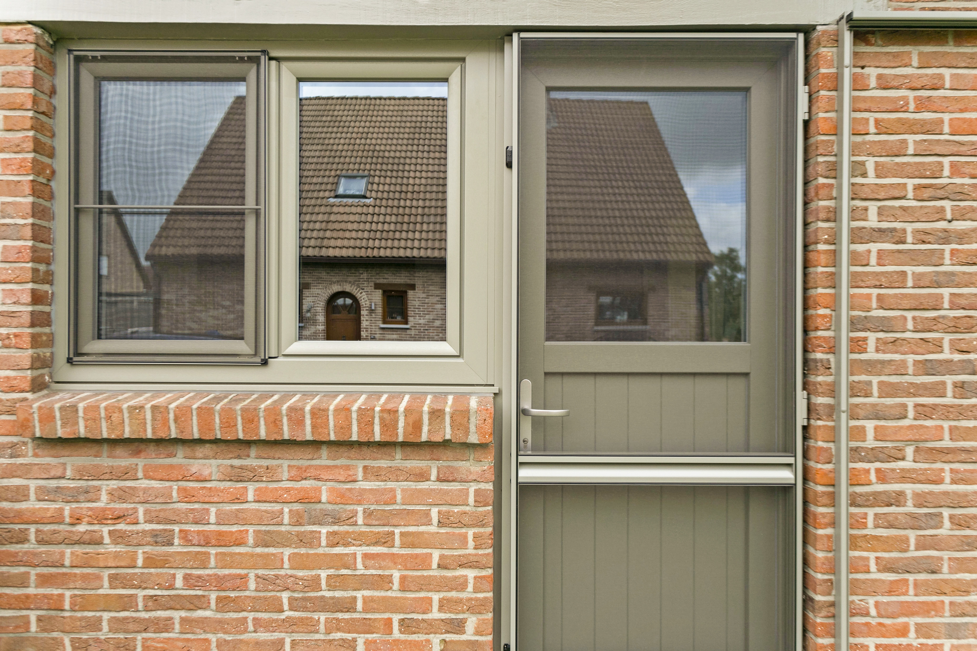 Betongrijs PVC-dubbel raam Impakt PLUS met draai-kipfunctie en vliegenraam, naast een semi-beglaasde deur in een bakstenen gevel.