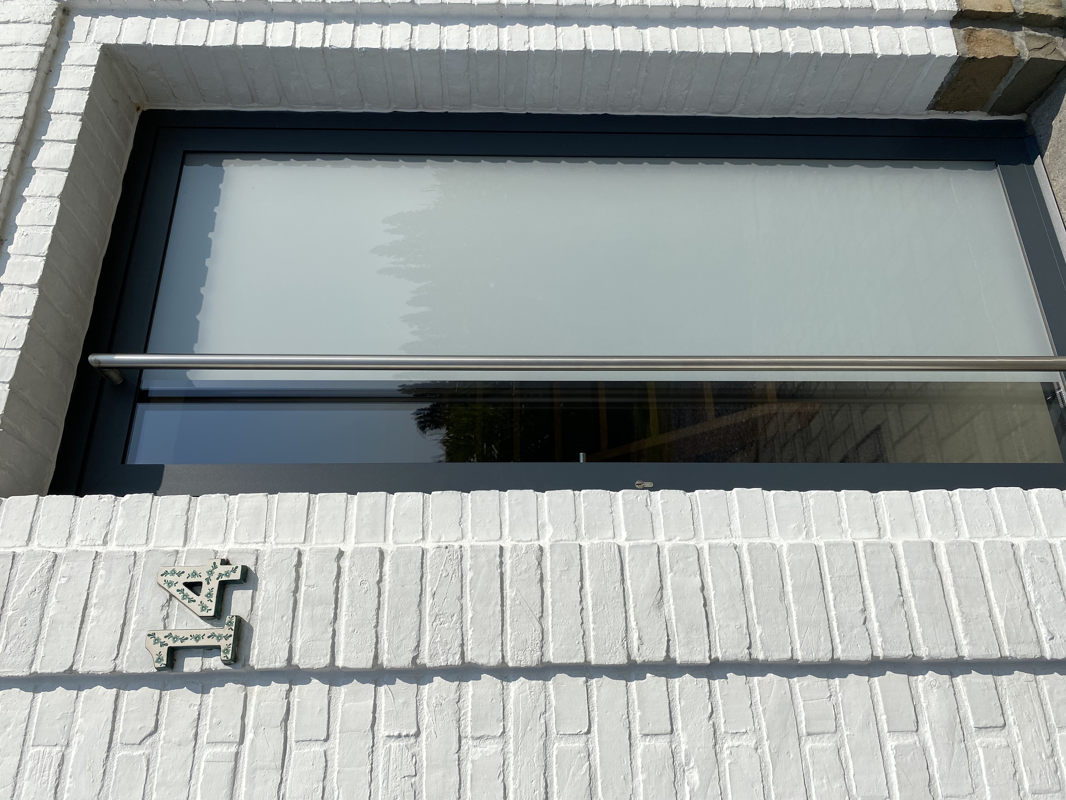 Glazen voordeur met donker raamprofiel in een witte bakstenen gevel van een vrijstaande woning, met mat glas en een lange verticale handgreep.