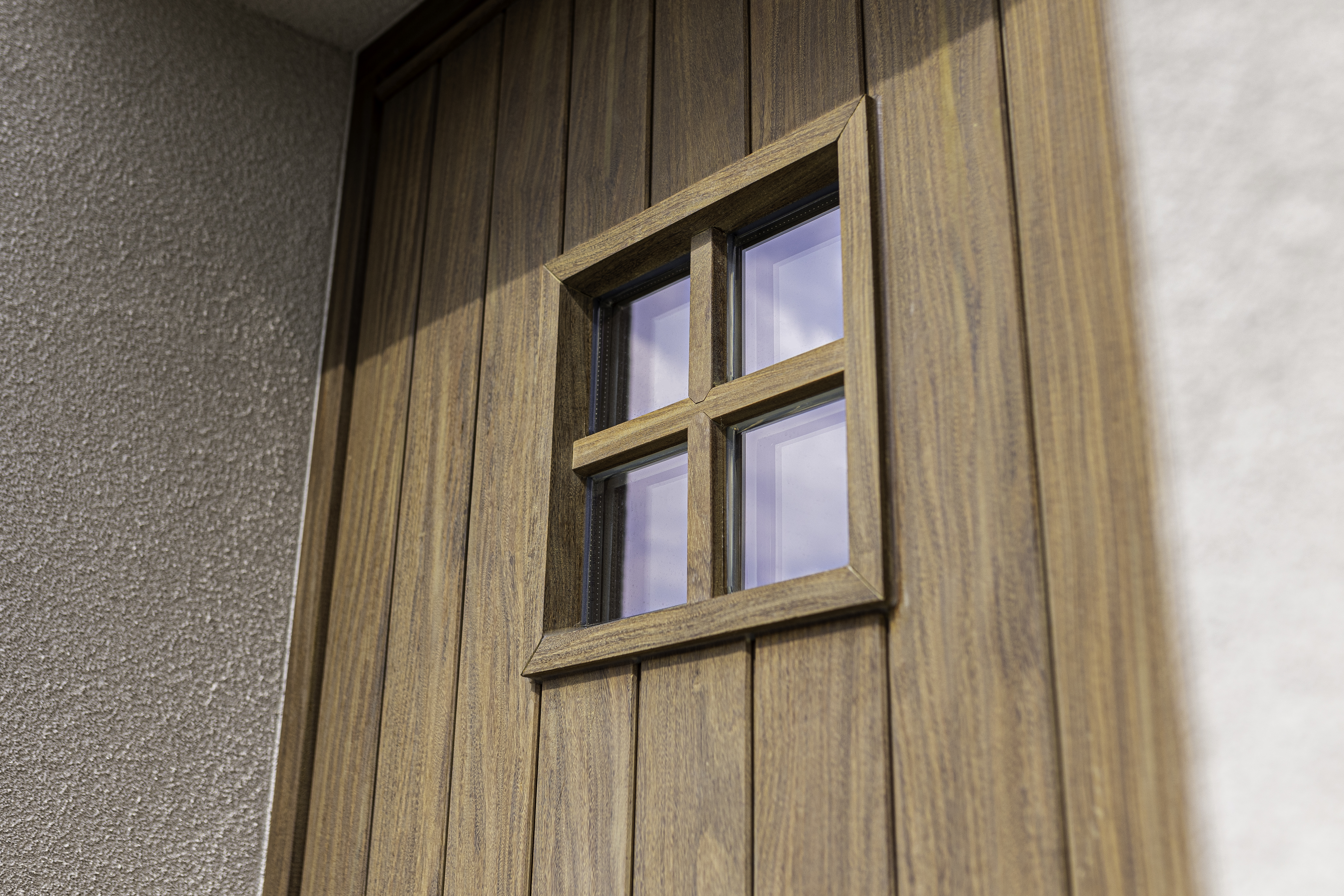 Gelakte voordeur in Afrormosia aan de gevel van een vrijstaande woning, met een klein raam met vier ruiten en zichtbare houtnerf.