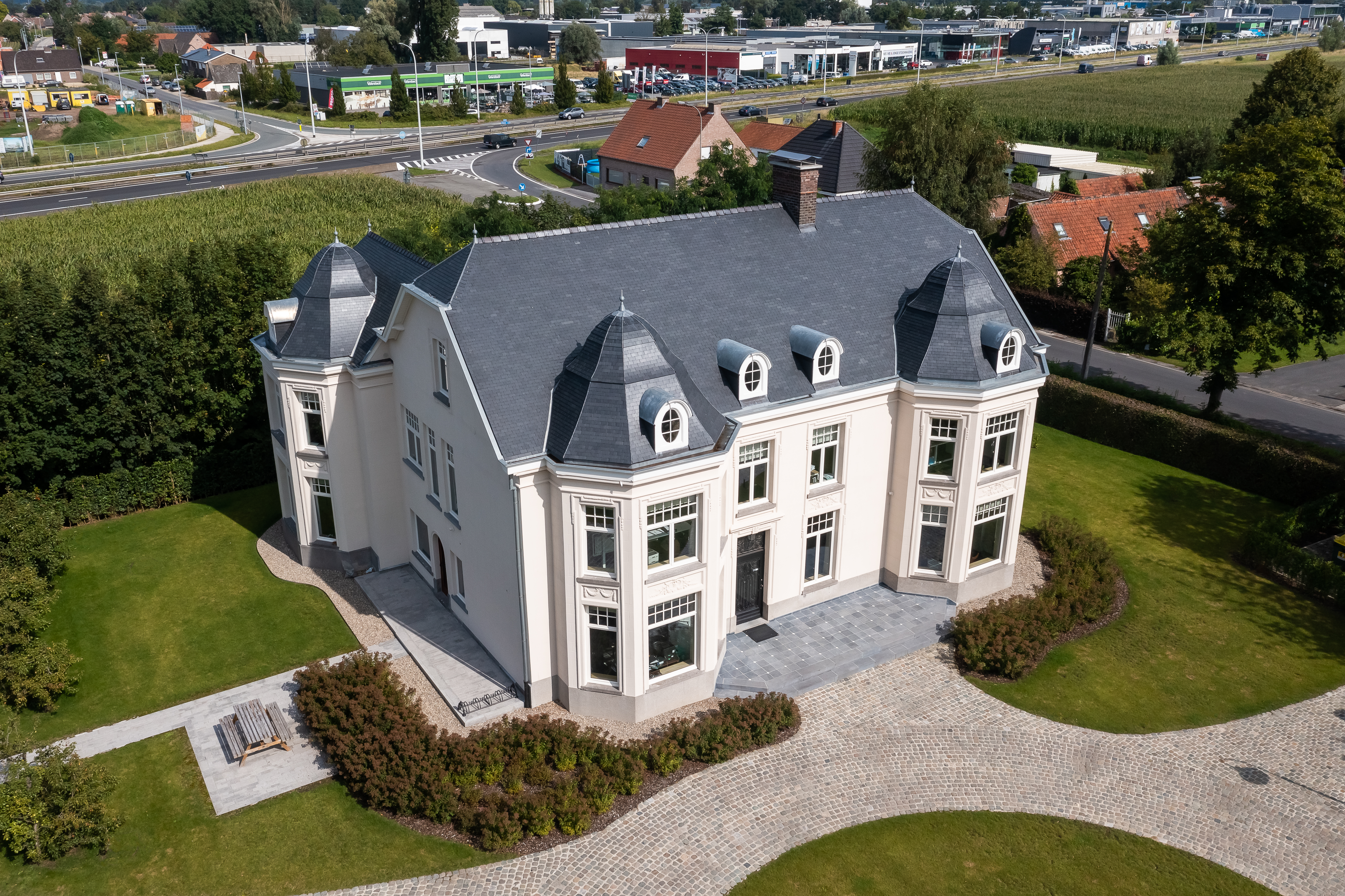 Gerenoveerde open bebouwing in verfijnde klassieke stijl, met een gevel in 9010TC Zuiver Wit, een donker leistenen dak, dakvensters en hoge witte ramen, omgeven door een tuin en grindpaden.