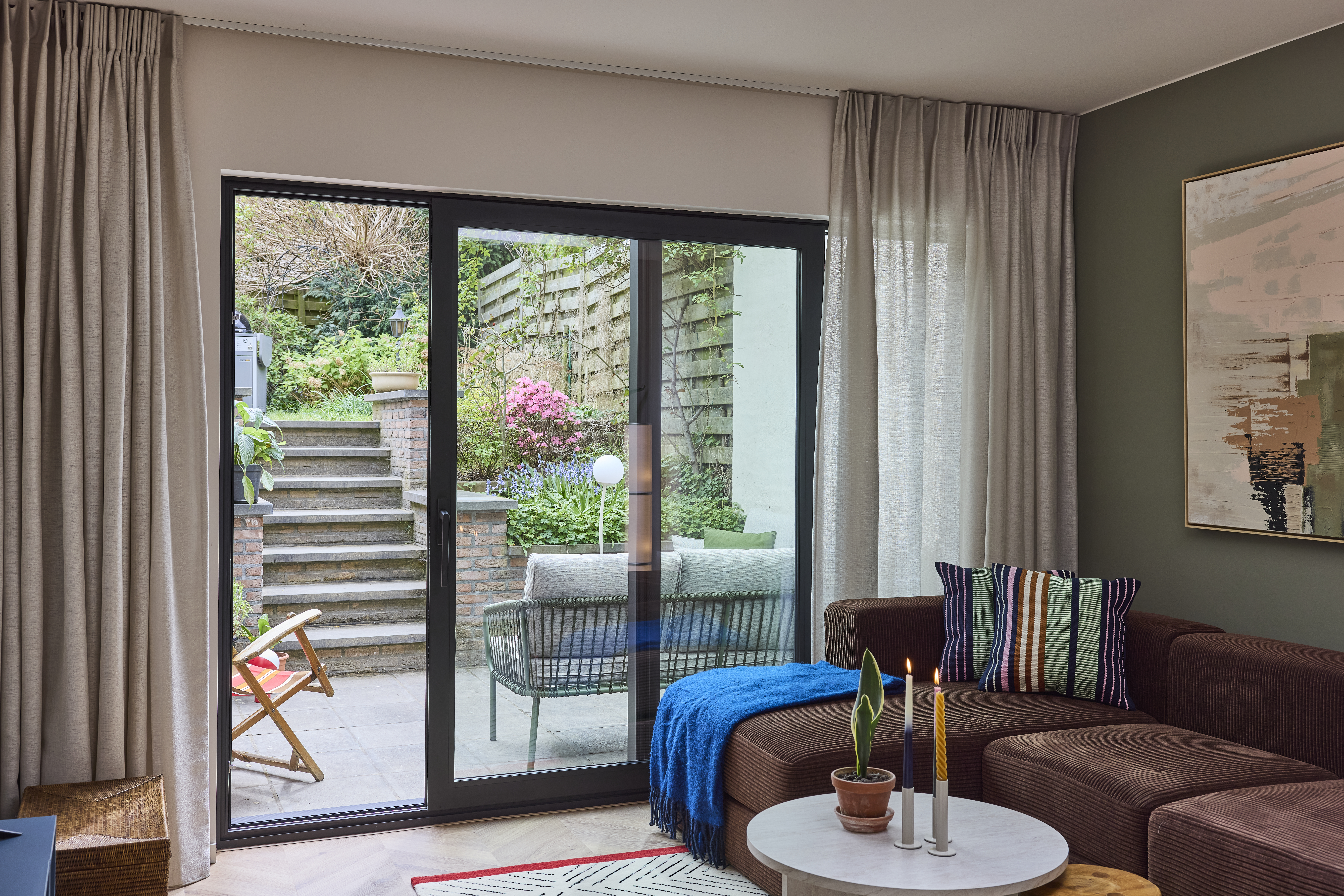Zwart aluminium ALU Visoglide Monorail-schuifraam in de woonkamer, met zicht op een groen terras en een buitentrap.