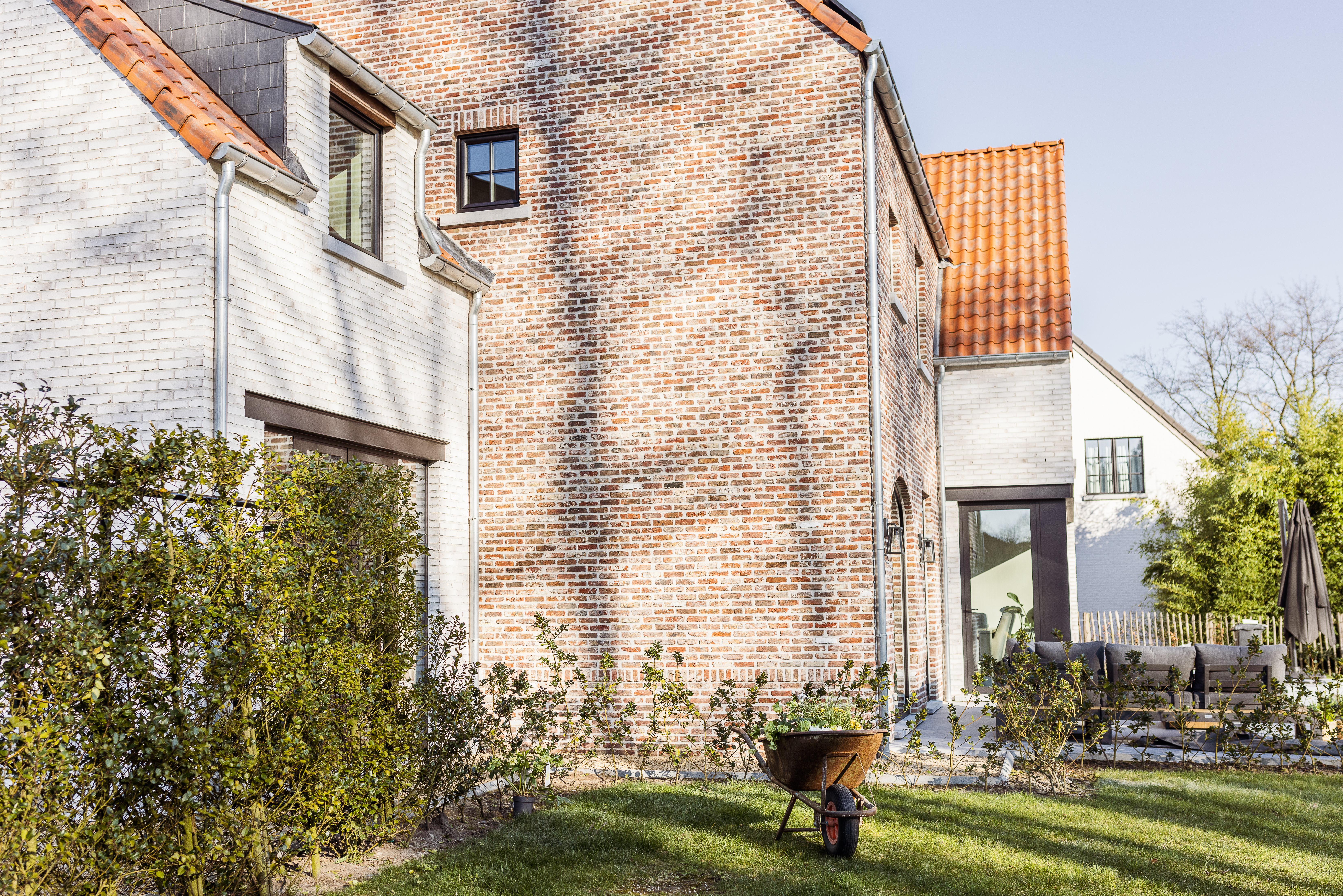 Bakstenen gevel met matzwarte raamprofielen en grote glaspartijen, naast een witte woning, met tuin en kruiwagen op de voorgrond.