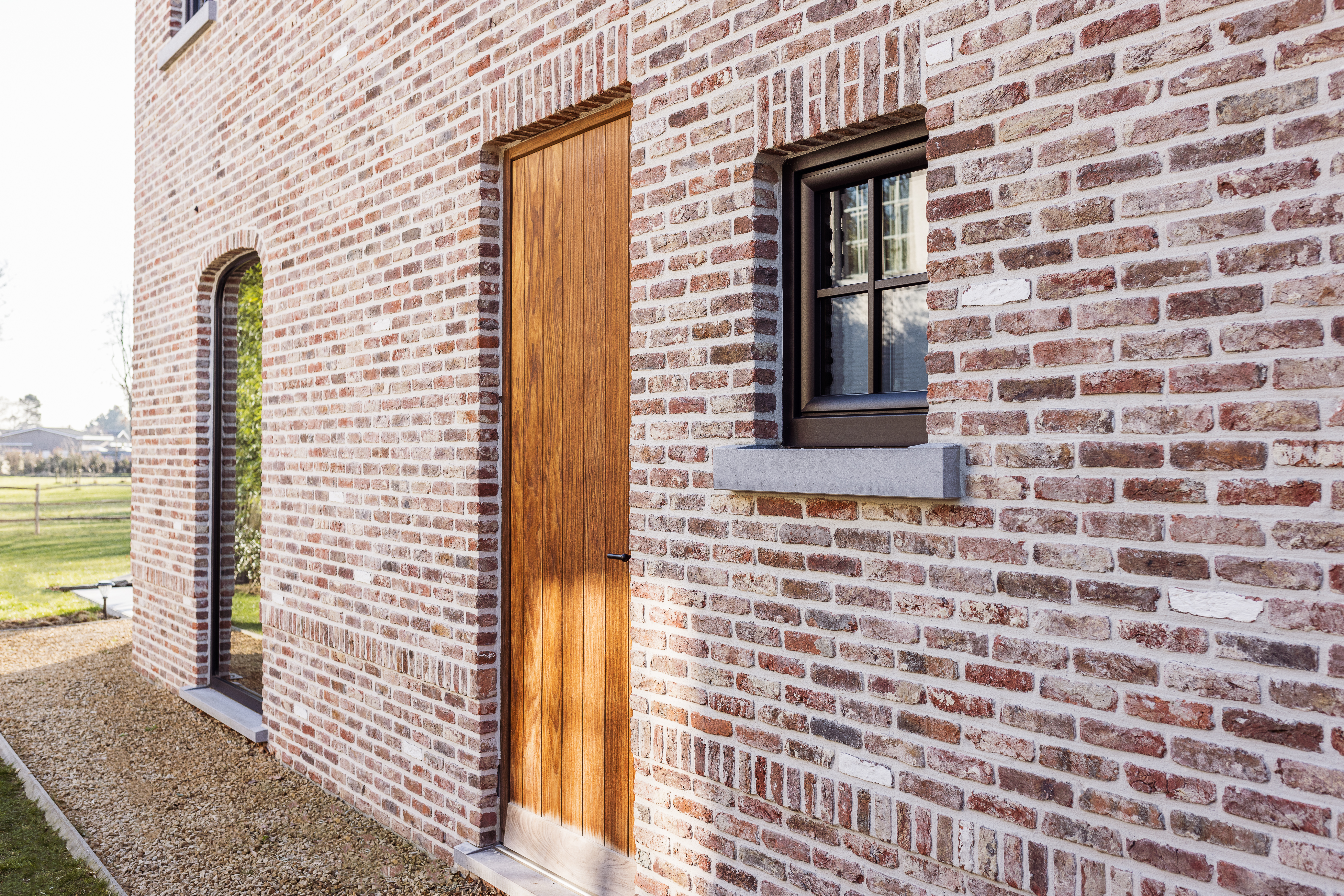 Bakstenen gevel met houten deur en klein raam met matzwart raamprofiel, naast een smalle glazen opening met zicht op het landelijke landschap.