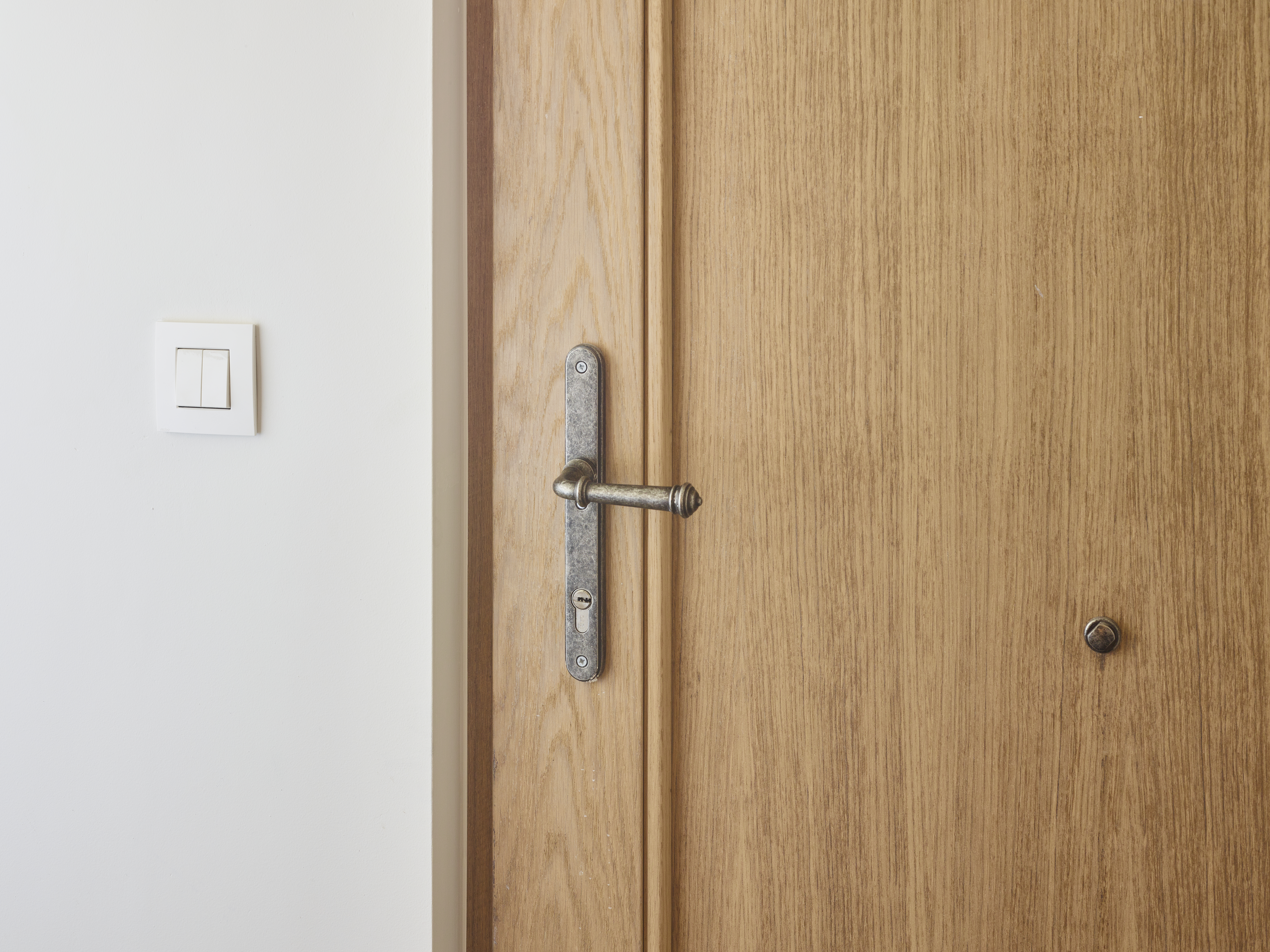 Afgelakt houten paneel in lichte eik met warme, landelijke uitstraling, naast een witte muur met lichtschakelaar en metalen beslag.