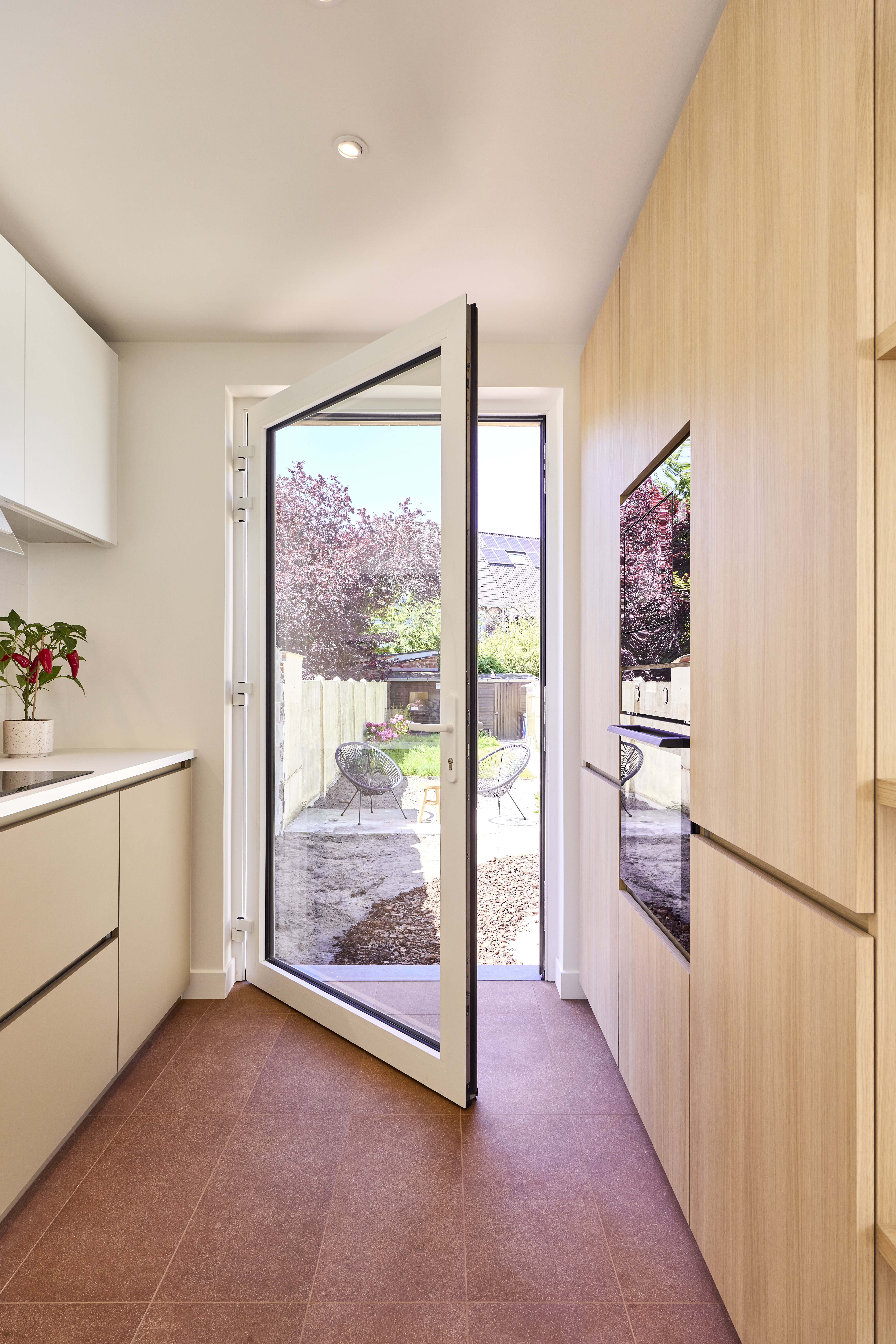Openstaande beglaasde aluminiumdeur in verkeerswit geeft vanuit een lichte ruimte toegang tot een terras en tuin; rechts staan hoge houten kasten.